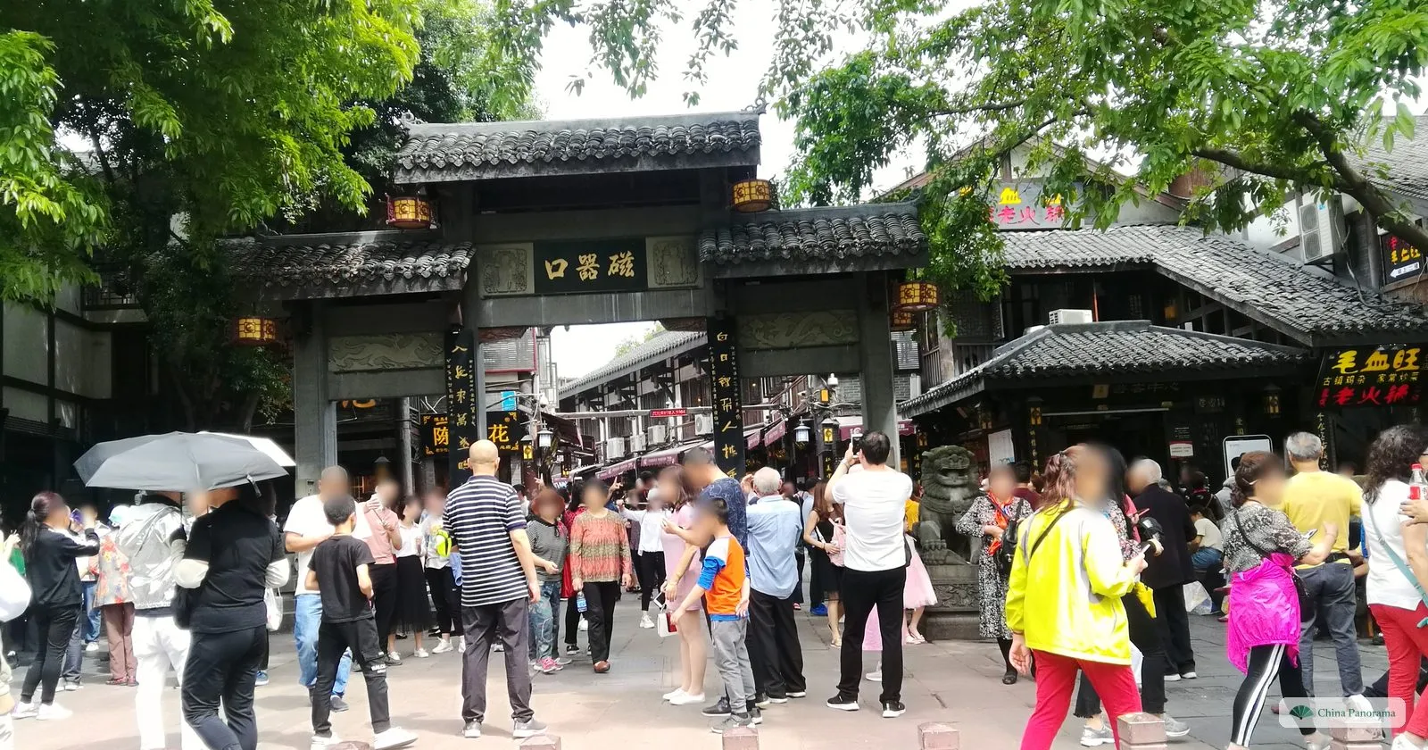 A crowd of holidaymakers in front of the Ciqikou gate, some with umbrellas, the plaque 磁器口 above the entrance