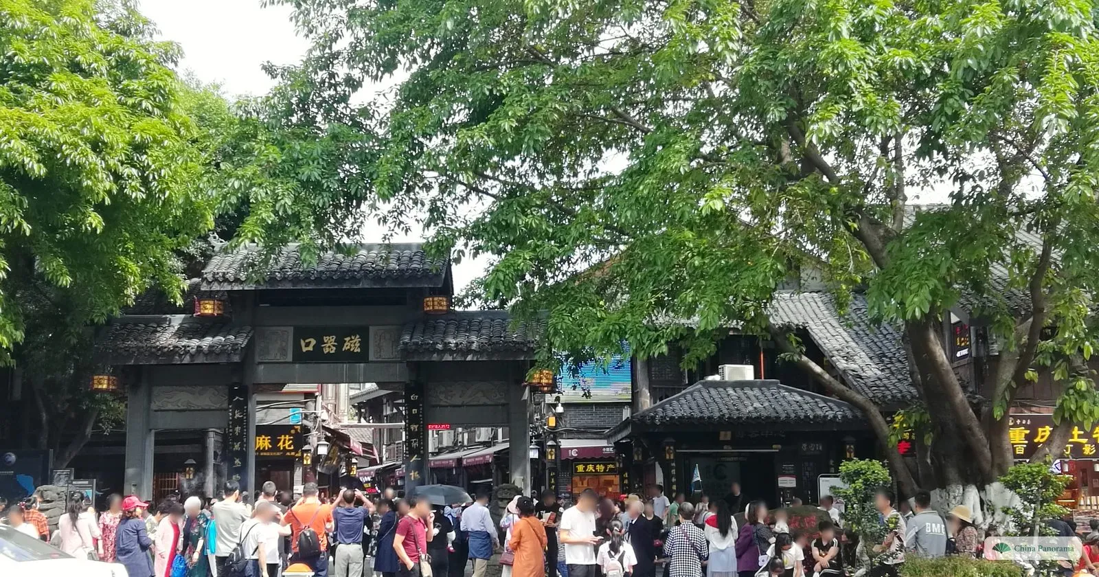 The grey-tiled main gate of Ciqikou with a black plaque 磁器口 under big trees, a dense crowd in front