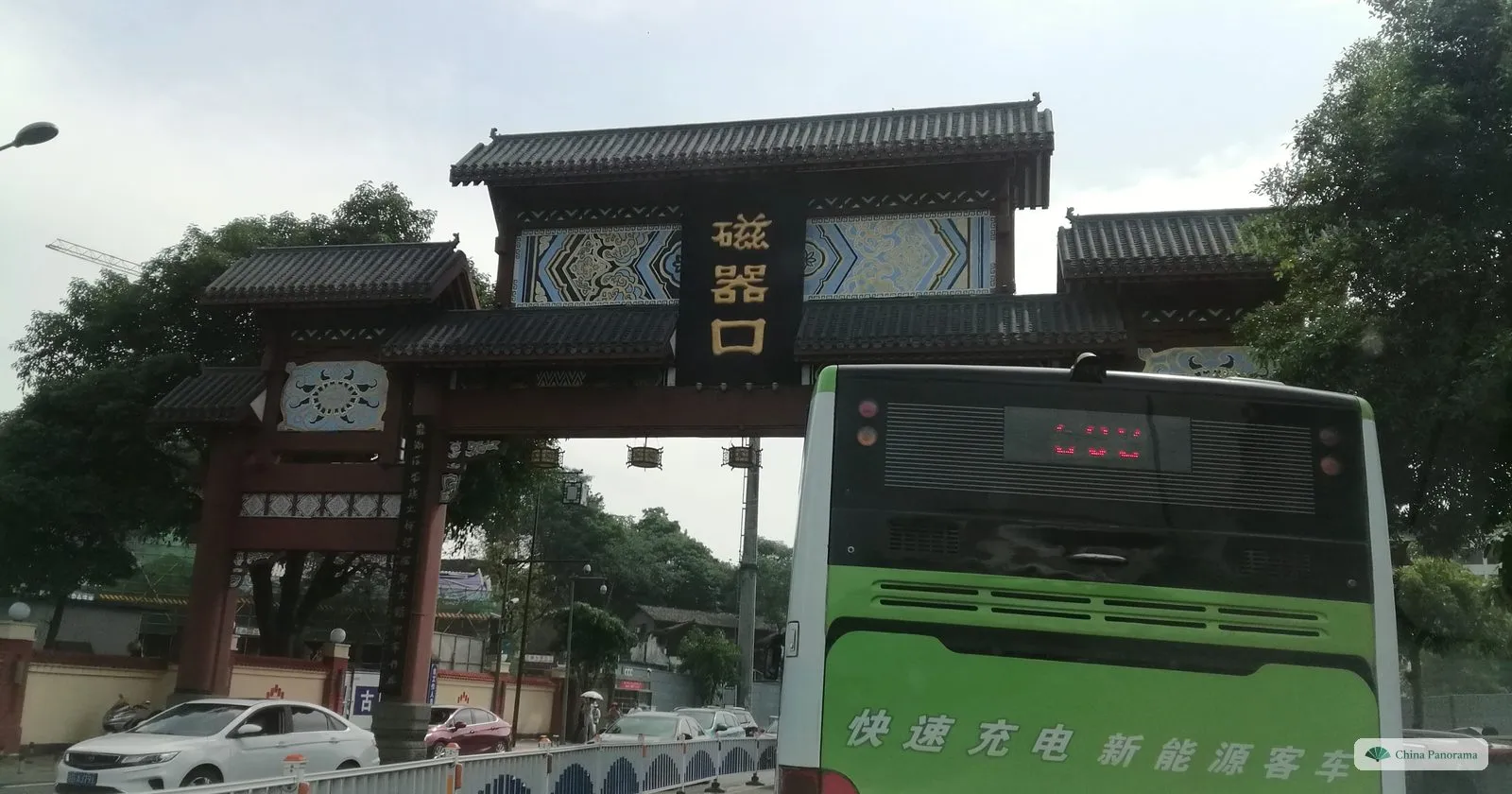 A carved wooden arch over the road reading 磁器口, with a green city bus passing underneath and trees around