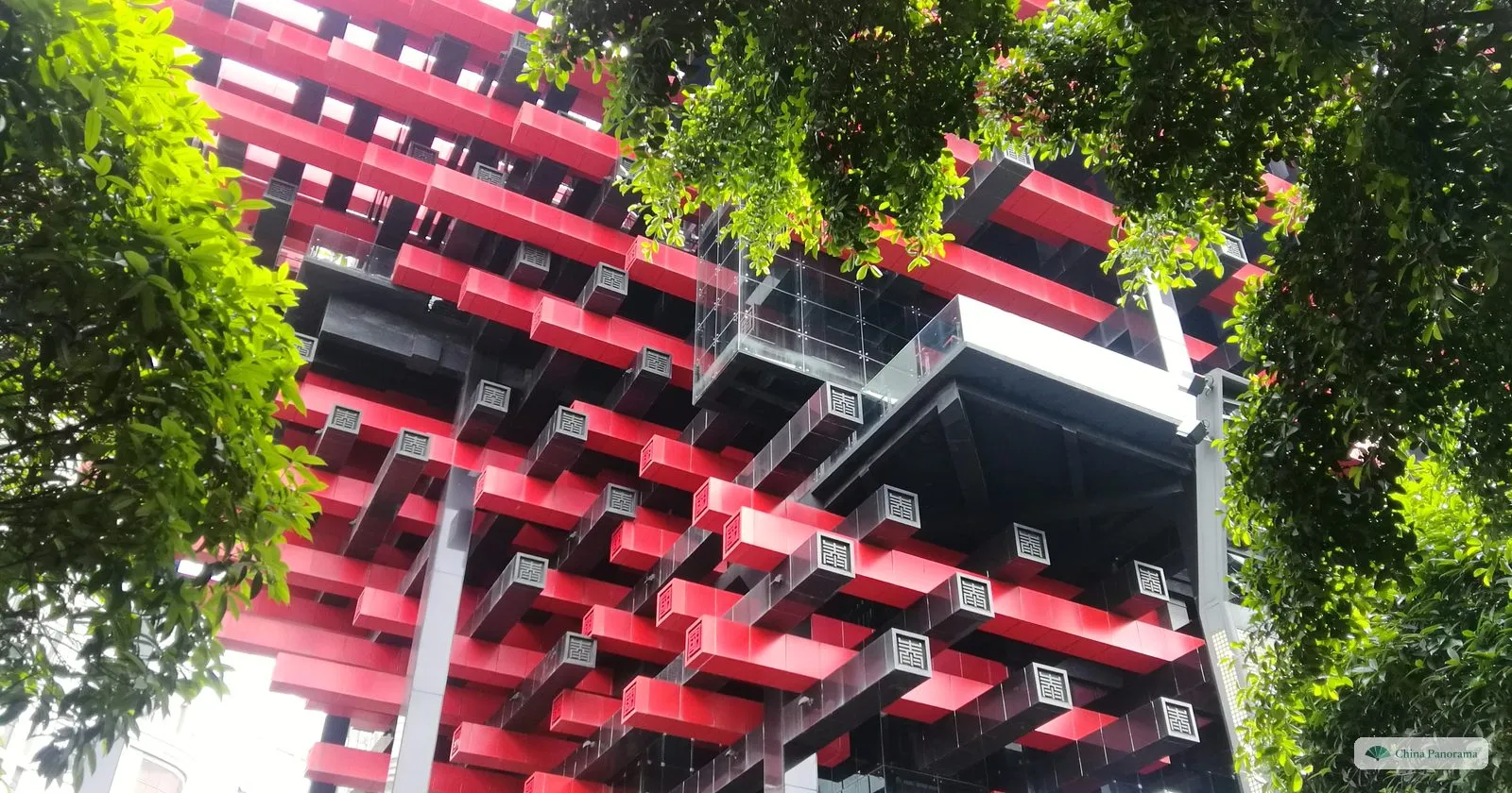 Looking up at the red and black beams of the Guotai Arts Center, stacked and cantilevered, framed by green branches