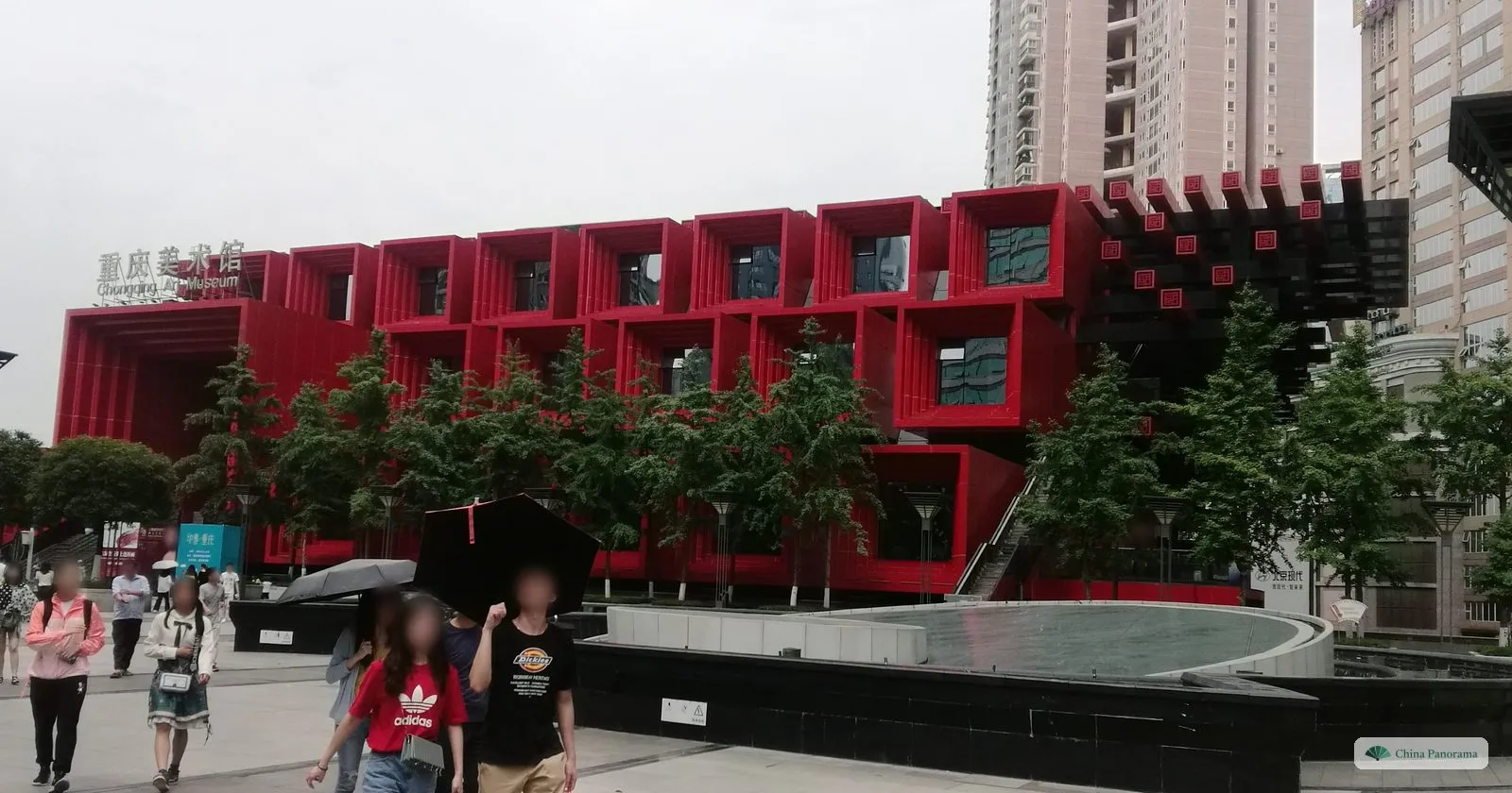 The Guotai Arts Center: a block of interlocking red and black beams sticking out in all directions, young trees and people with umbrellas in front