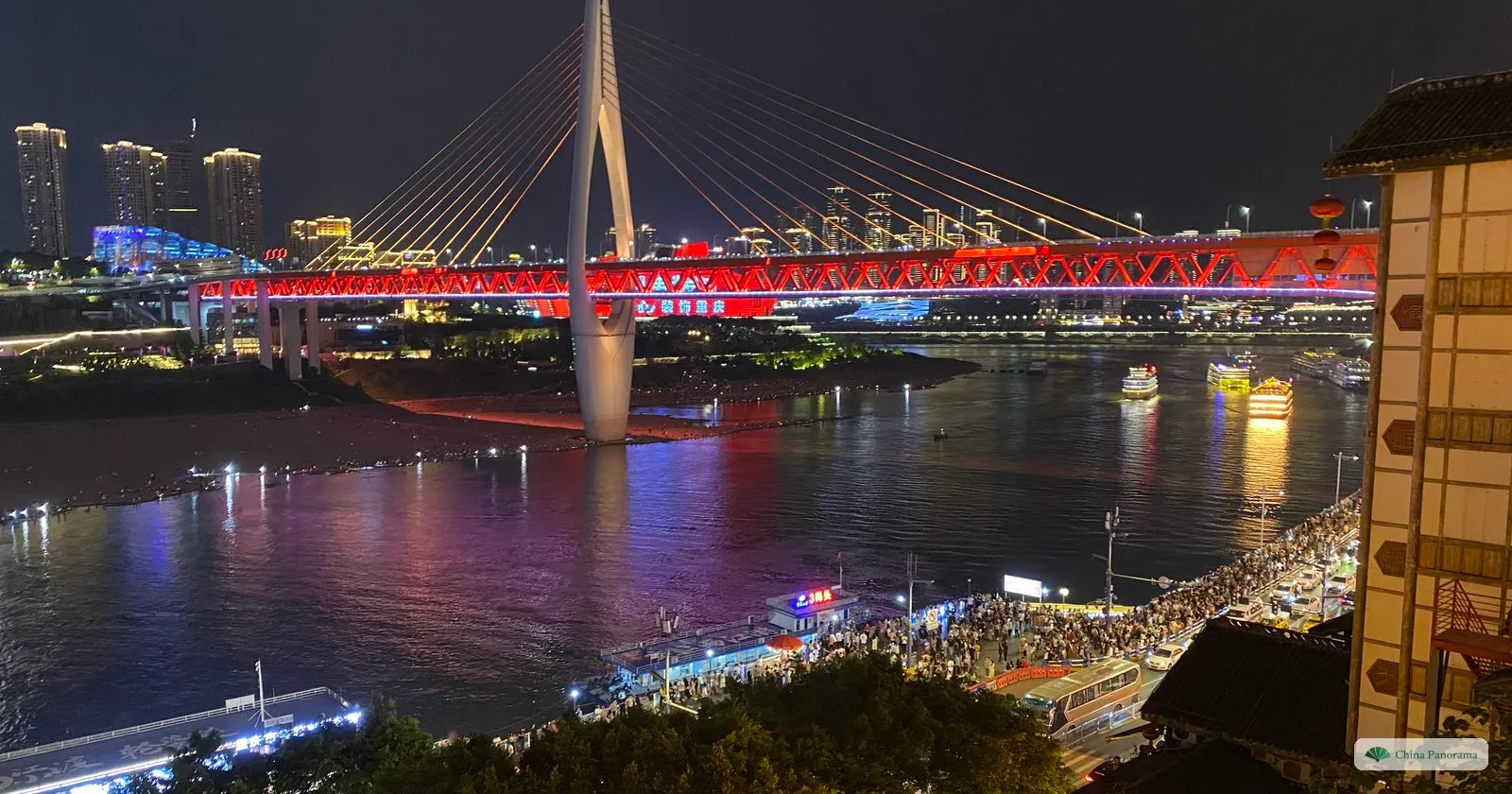 The Qiansimen Bridge at night from a terrace of Hongyadong: the red-lit truss and white tower over the dark river, crowds on the riverside road below