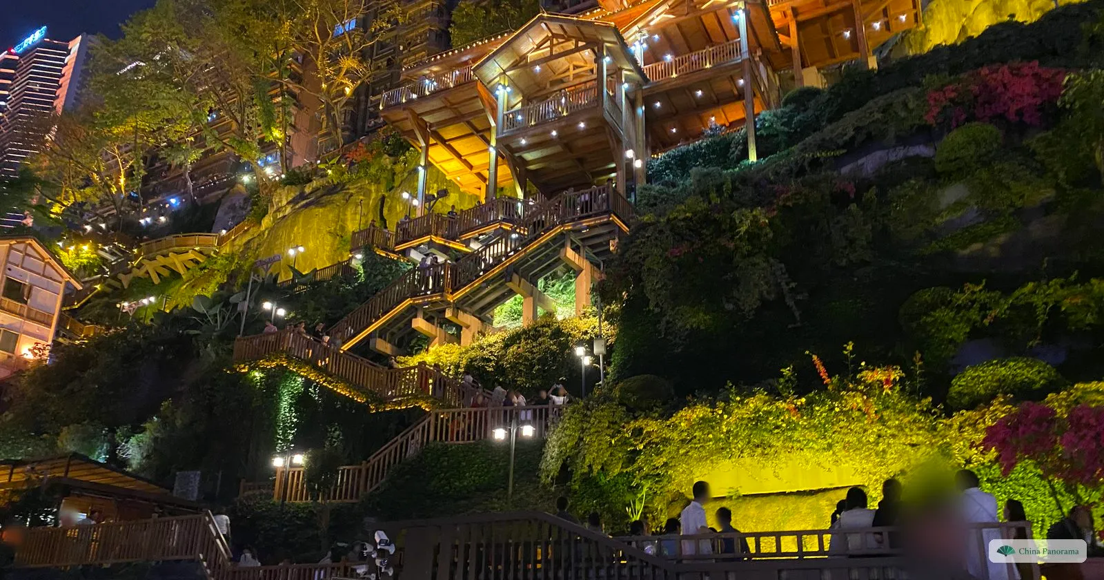 Wooden stairways and terraces climbing the cliff beside Hongyadong at night, lit yellow among dense green plants, with a few people at a railing below