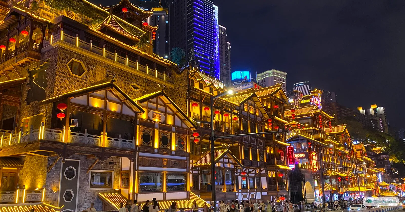 The lit front of Hongyadong seen along the riverside road at night, with a white car passing, a railing in front and a blue-lit office tower behind the eaves