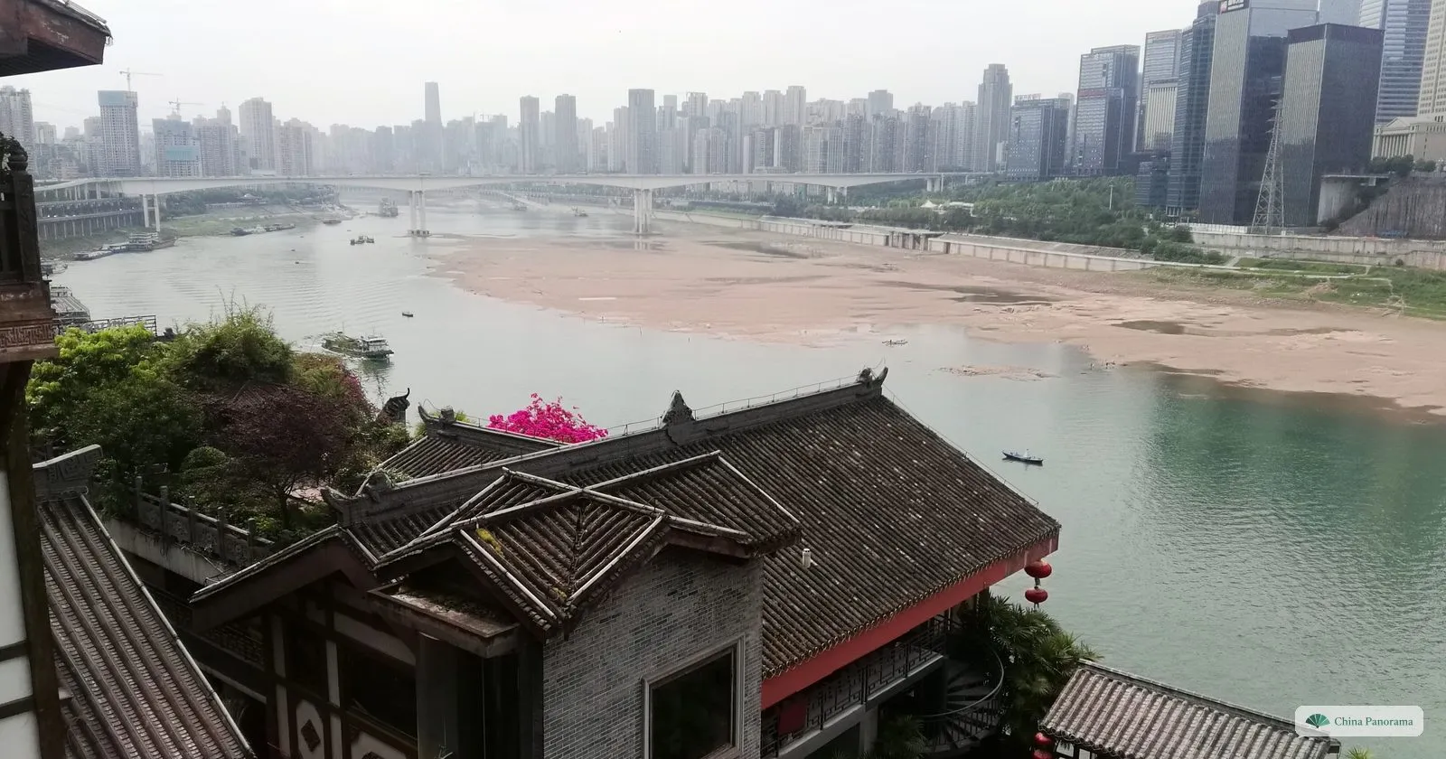 View from the roofs of Hongyadong over the green Jialing River, with wide sandbanks at low water, a pink bougainvillea and the towers of the north bank in haze