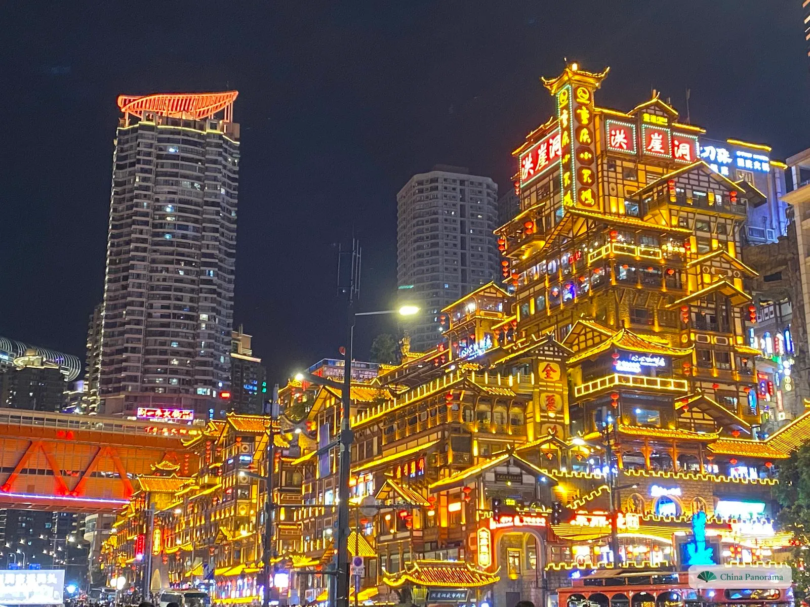 Hongyadong at night: tiers of wooden balconies, upturned eaves and red lanterns outlined in gold light, with the red characters 洪崖洞 on the top, a residential tower to the left and the red truss of the Qiansimen Bridge at the edge