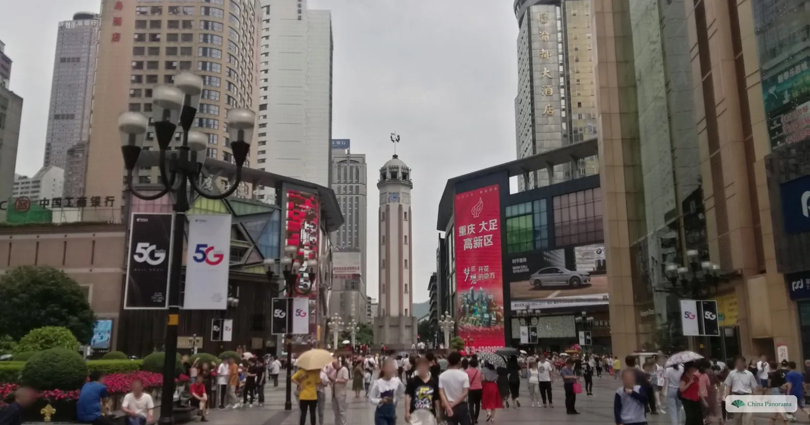 The Jiefangbei clock tower at the end of a crowded pedestrian street, between department stores with red banners and street lamps, on a grey day
