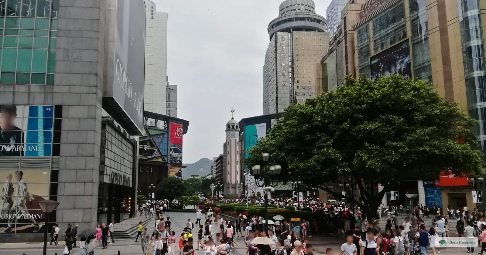 A broad pedestrian street in Jiefangbei under a big banyan tree, crowded with shoppers, the clock tower small in the distance between glass towers