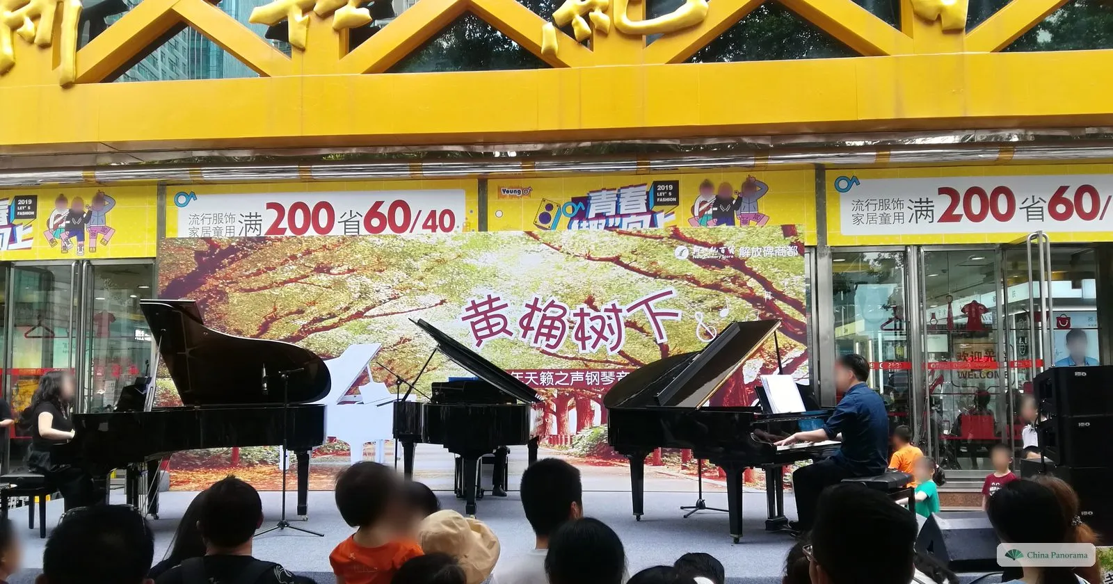 Four black grand pianos on an open-air stage under the yellow arch of a department store, with a banner reading 黄桷树下 and a crowd watching