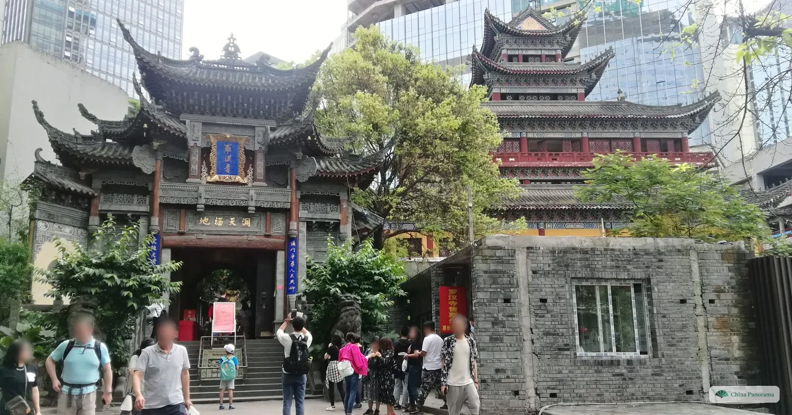 The gate of Luohan Temple with curved, layered eaves and a blue plaque, a red pagoda-like hall behind, an old brick wall and visitors on the steps