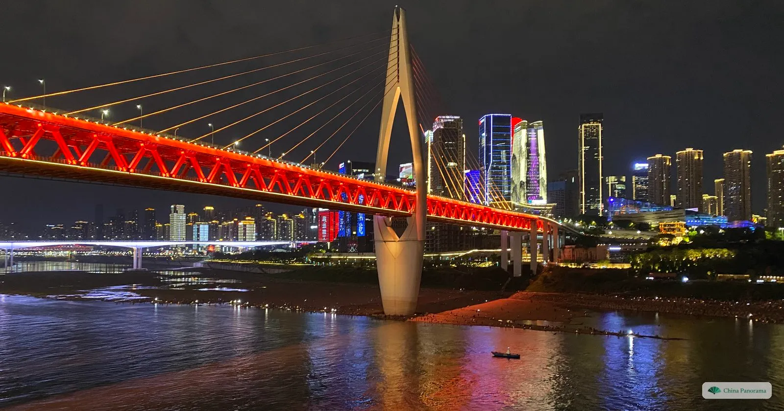 The Qiansimen Bridge at night from the riverbank: a white A-shaped tower with fanned cables and a red-lit steel truss over the river, lit towers beyond