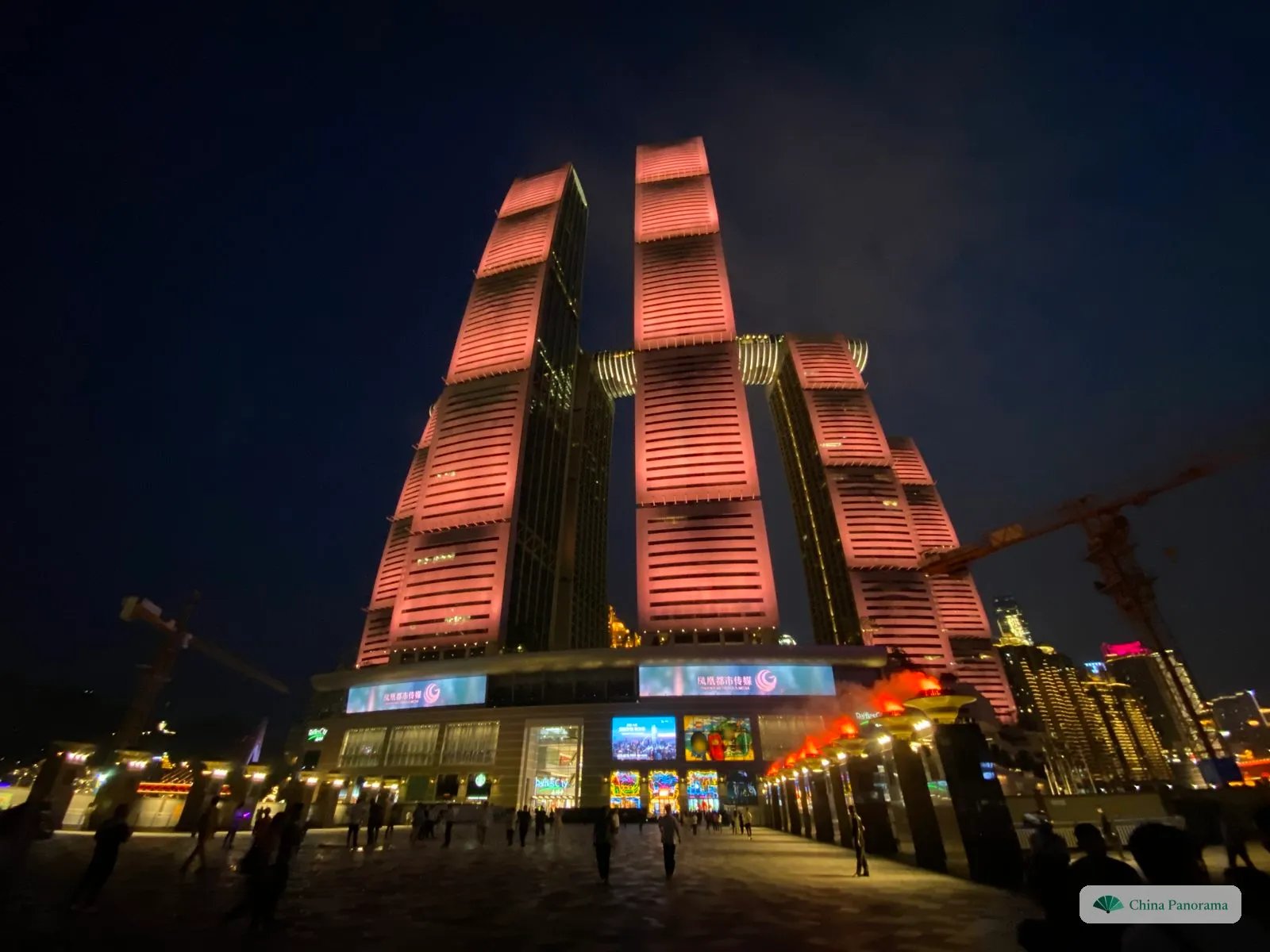 Raffles City Chongqing at night from the plaza: tall towers lit pink with a horizontal skyway joining them high up, a lit mall at the base and people walking in front