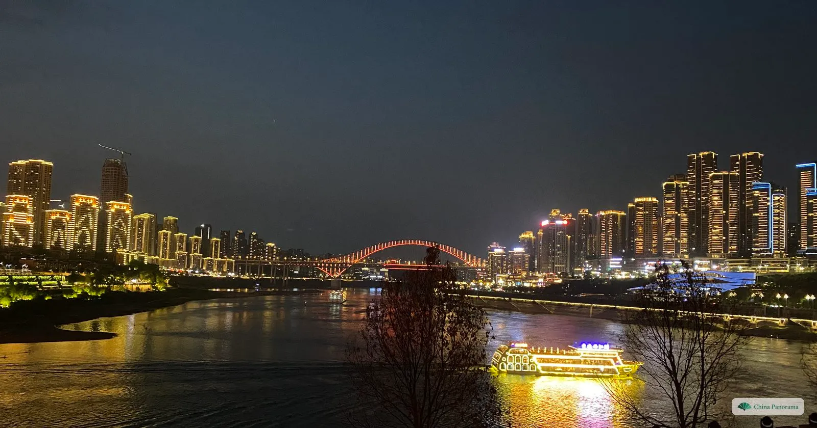 The Yangtze at night: the lit arch of a bridge in the distance between rows of illuminated towers, a glowing tour boat and bare trees on the near bank