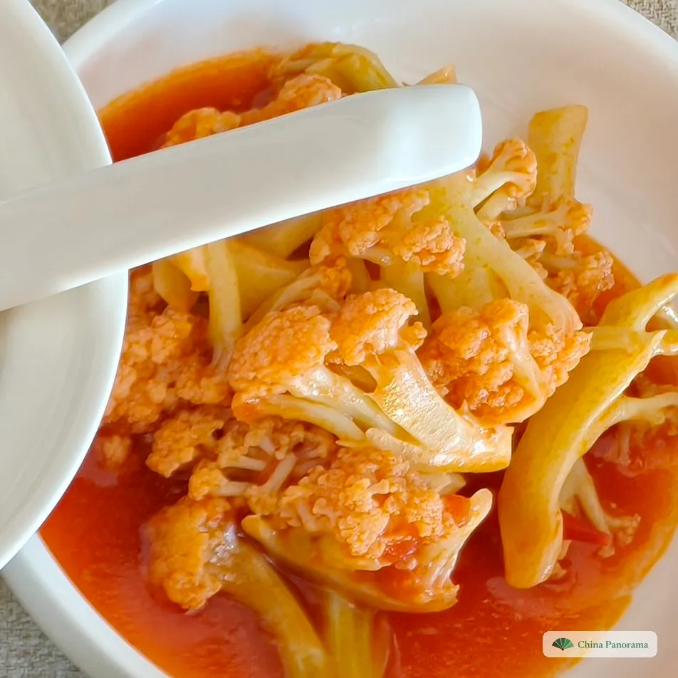 A white bowl of cauliflower florets and stems in red tomato sauce, with a white spoon