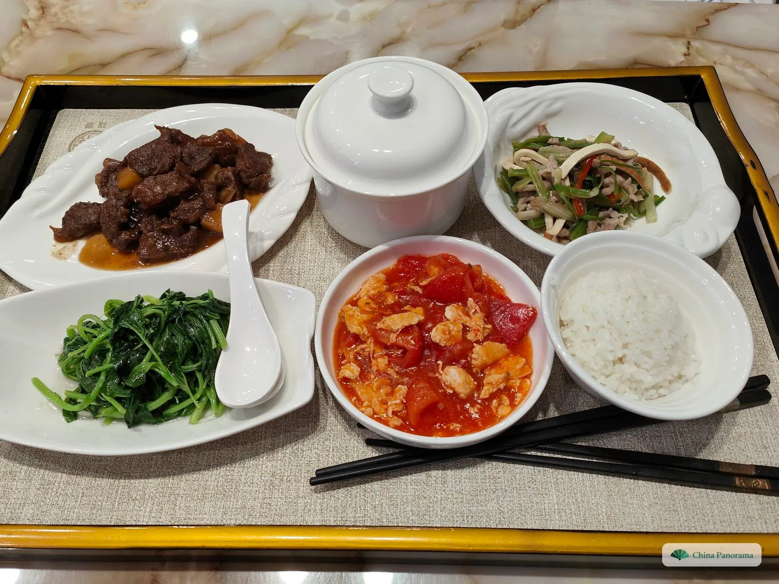 A set lunch on a tray from above: braised beef, a lidded white soup pot, stir-fried celery with tofu strips, pea shoots next to a spoon, a round bowl of tomato and egg in red sauce, and a bowl of rice with chopsticks