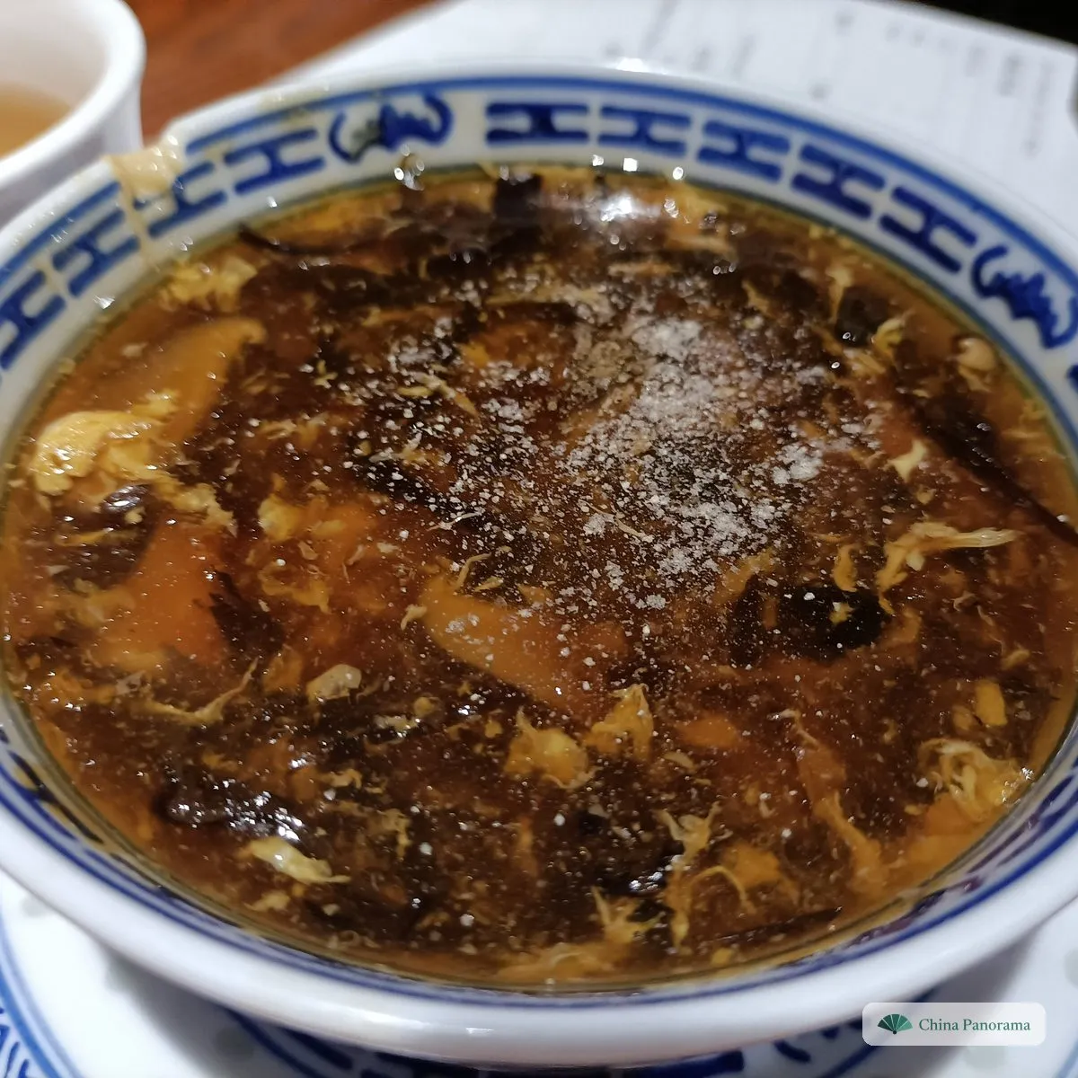 A blue and white bowl of thick dark-brown soup with egg, tofu strips and black wood ear, sprinkled with white pepper