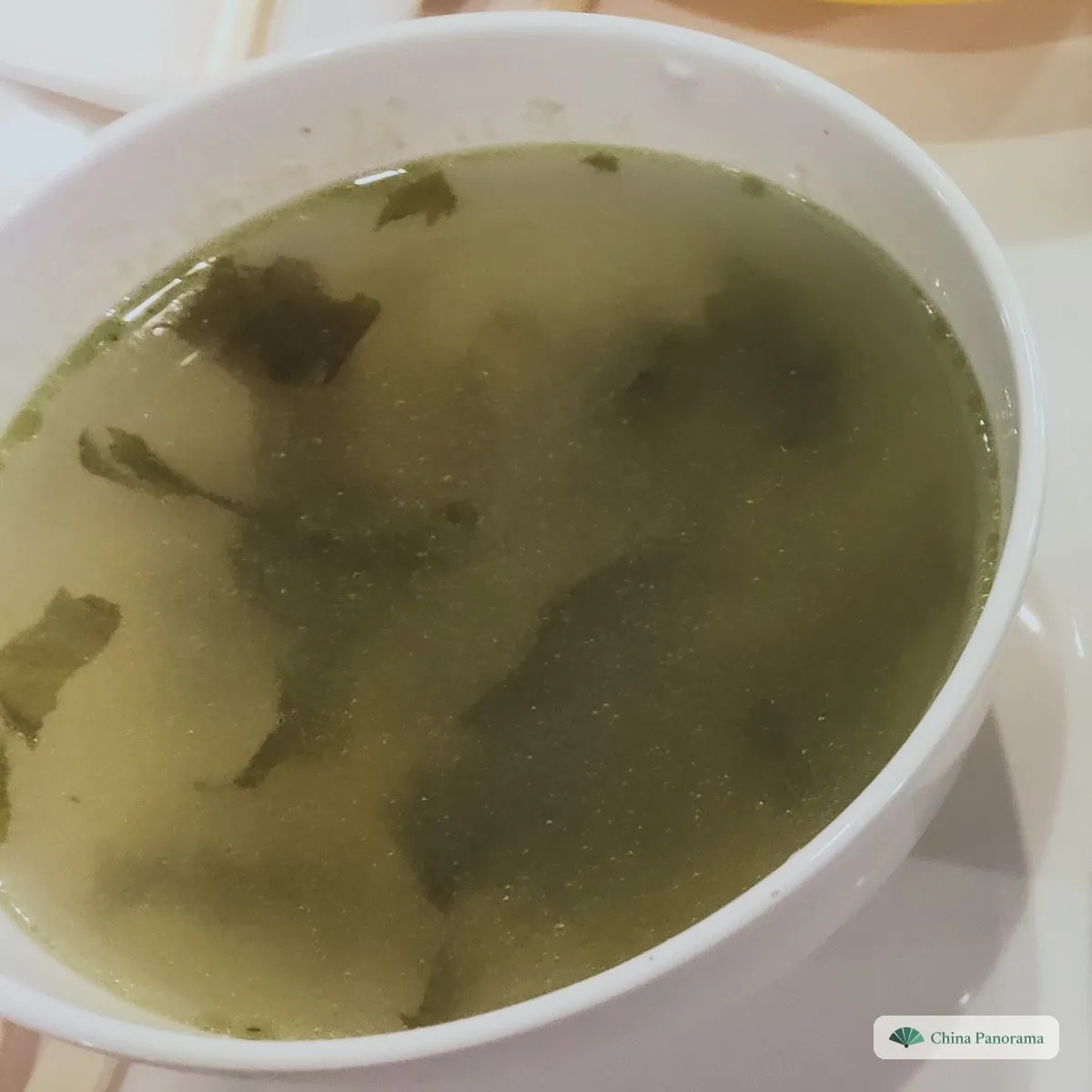 A white bowl of pale green broth with dark flakes of seaweed floating in it