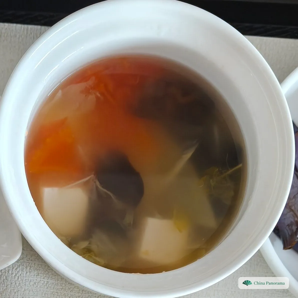 A white lidded soup pot with clear broth, pieces of tomato, white tofu cubes and dark mushrooms