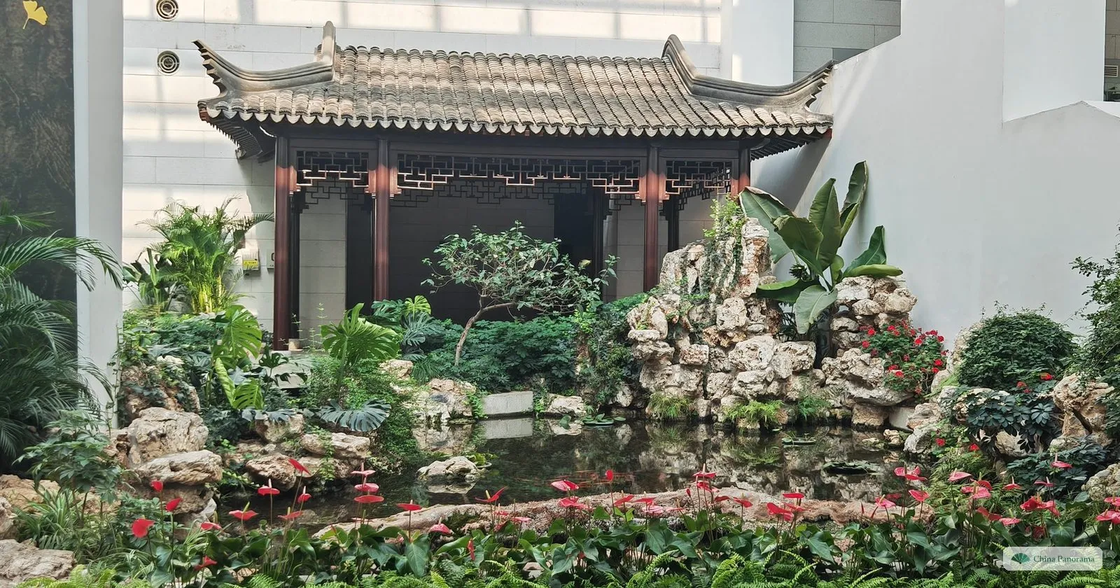 A grey-roofed pavilion above a rockery and pond with flowers, built inside the glass-roofed hall of the Museum of Chinese Gardens