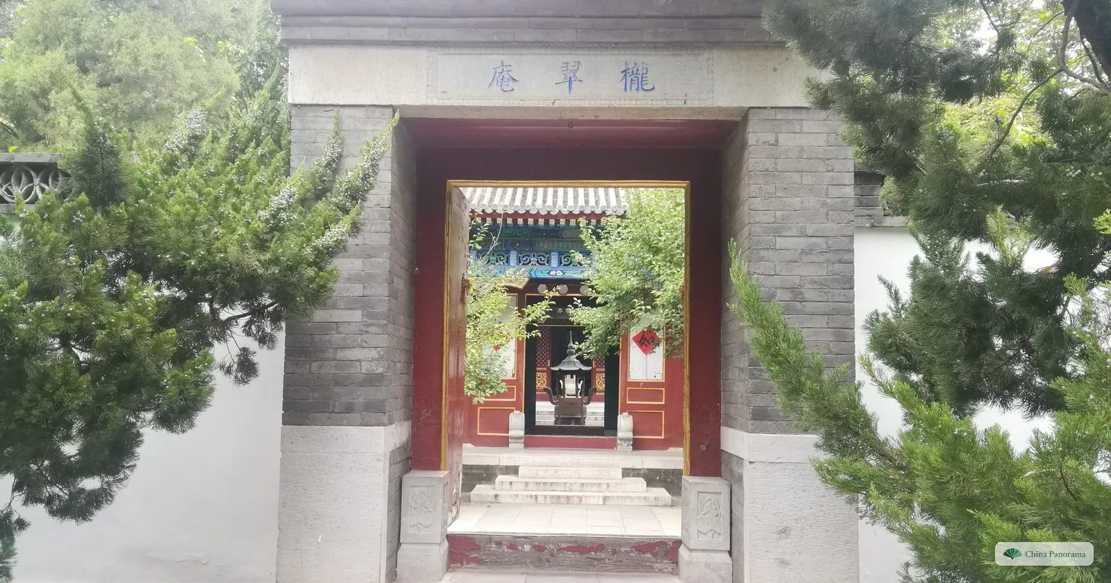 A grey brick gateway with a red door frame in Grand View Garden, looking through to a courtyard, with pines on both sides