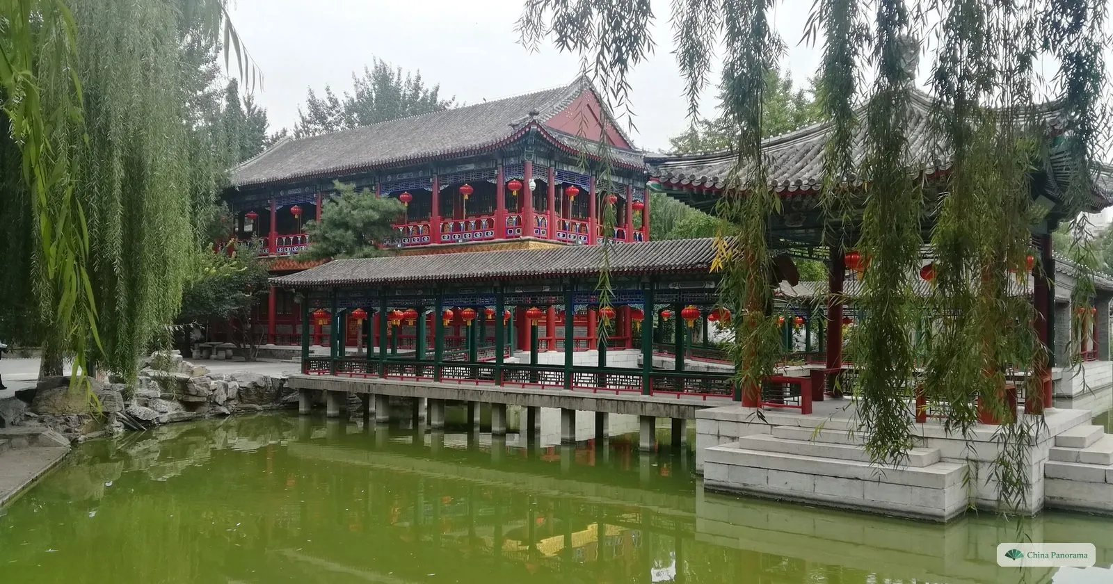 Waterside halls with red columns and grey roofs on a green pond in Grand View Garden, framed by weeping willows
