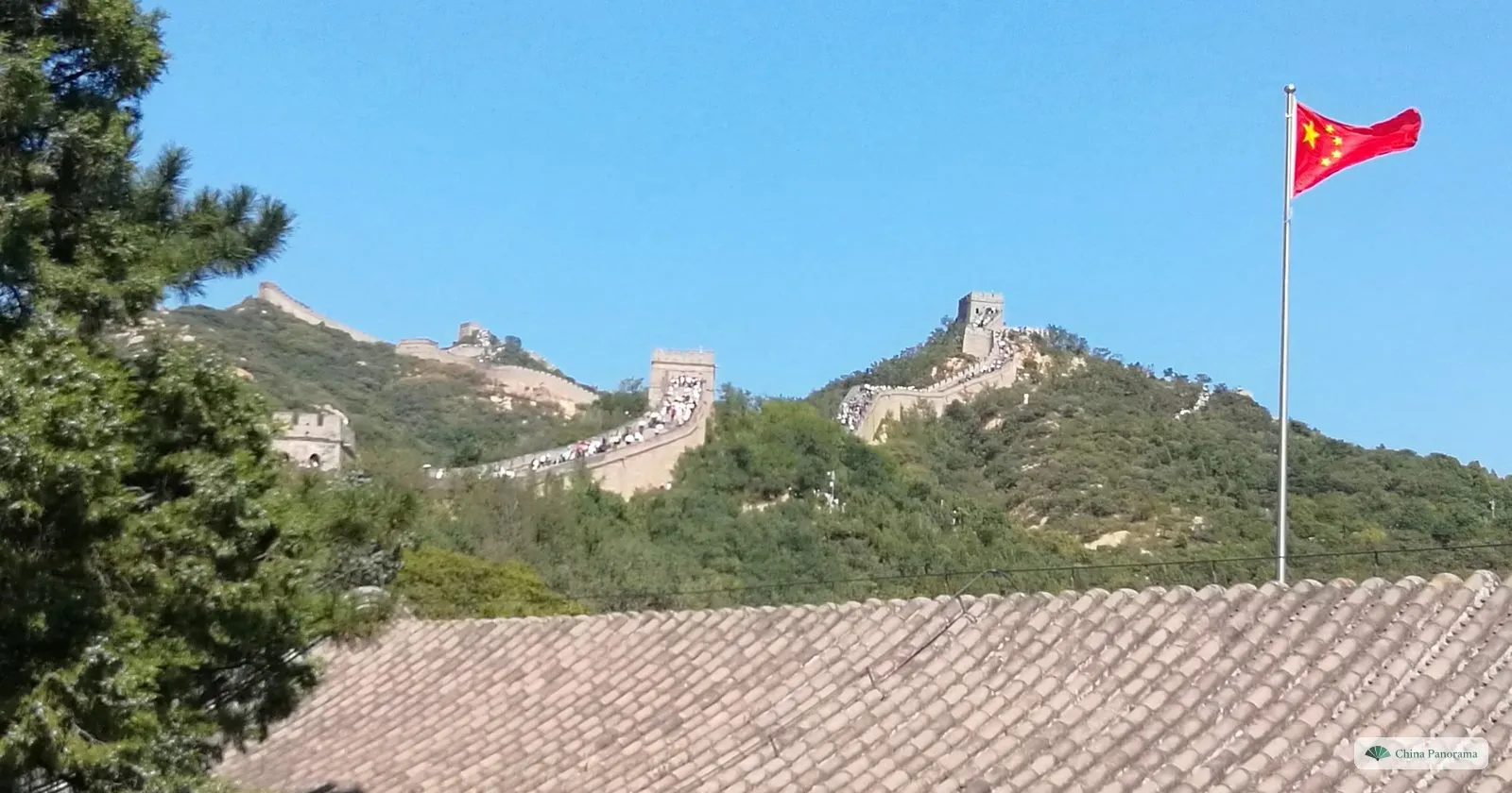 The Great Wall at Badaling crossing two wooded ridges with watchtowers, seen over a grey tiled roof, with a Chinese flag on a pole