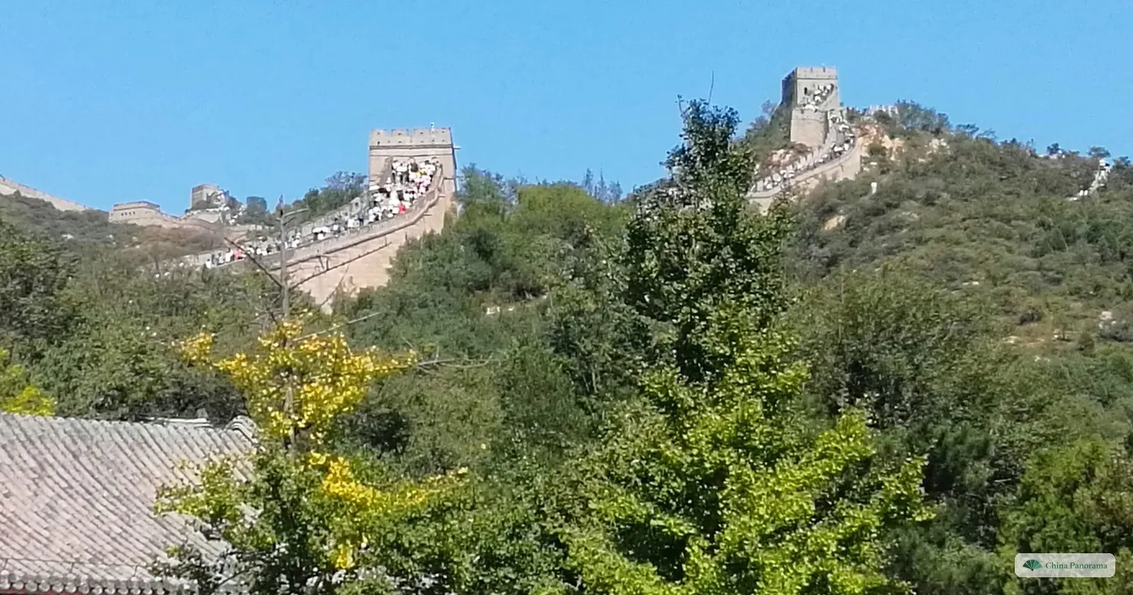 Two watchtowers of the Great Wall at Badaling on a hilltop, the wall between them crowded with visitors, above trees turning yellow