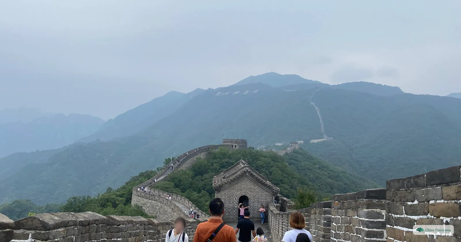 The Great Wall at Mutianyu on a hazy day: visitors walking down the wall towards a watchtower, with the wall continuing up a forested mountain and large white characters on the hillside