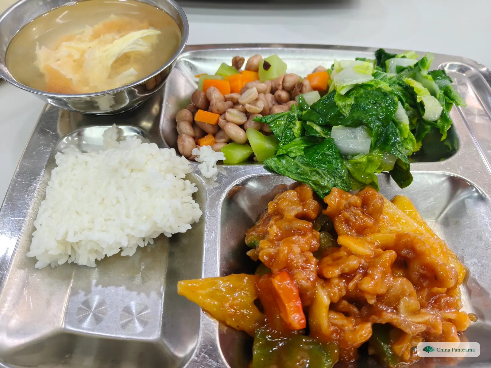 A steel canteen tray from above: sweet and sour pork with pineapple, green pepper and carrot in an orange sauce, a scoop of rice, peanuts with diced carrot and cucumber, lettuce, and a bowl of soup