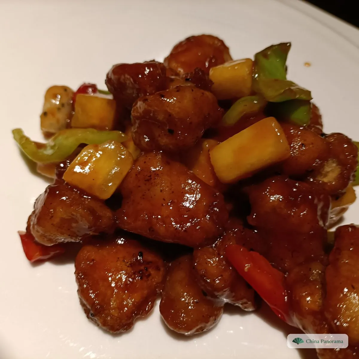 Bite-sized pieces of battered pork in a glossy red-brown sauce with chunks of pineapple and green and red pepper on a white plate