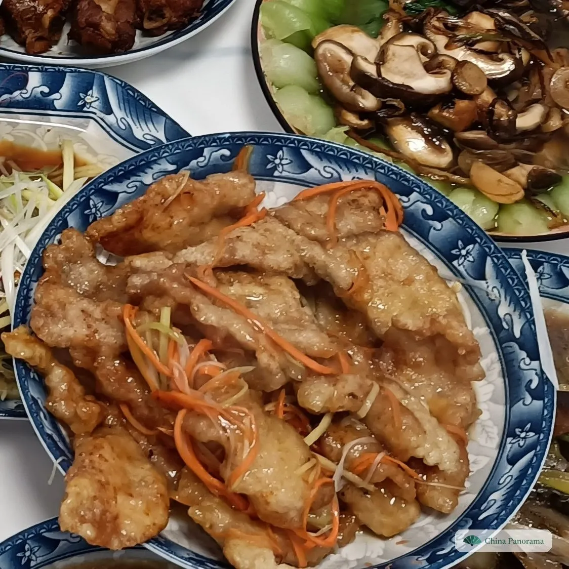 Large, flat slices of pale golden fried pork with shreds of carrot and scallion in a blue-and-white bowl, other dishes around it