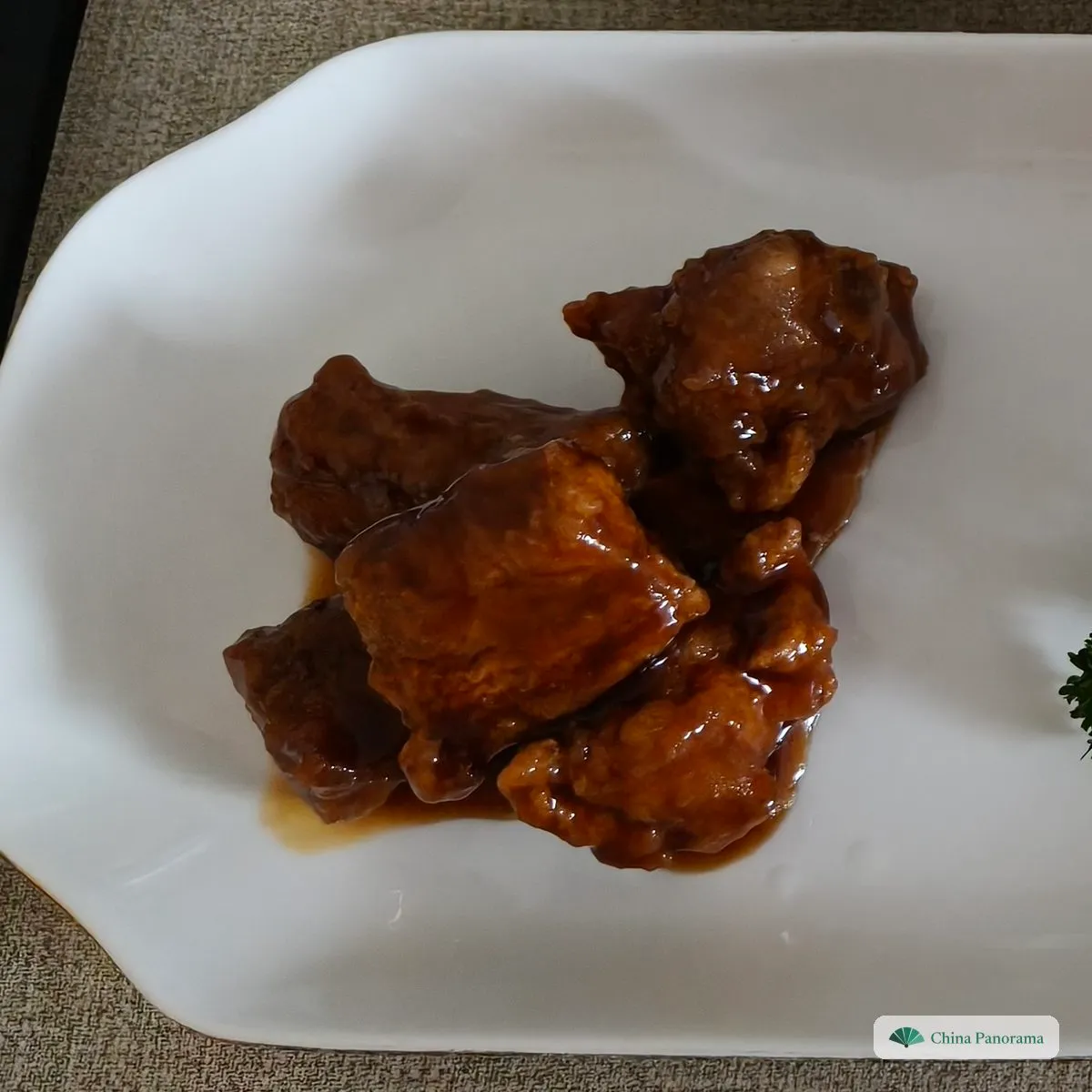 A few dark brown, glossy pork ribs in a little sauce on a white plate, with a sprig of parsley