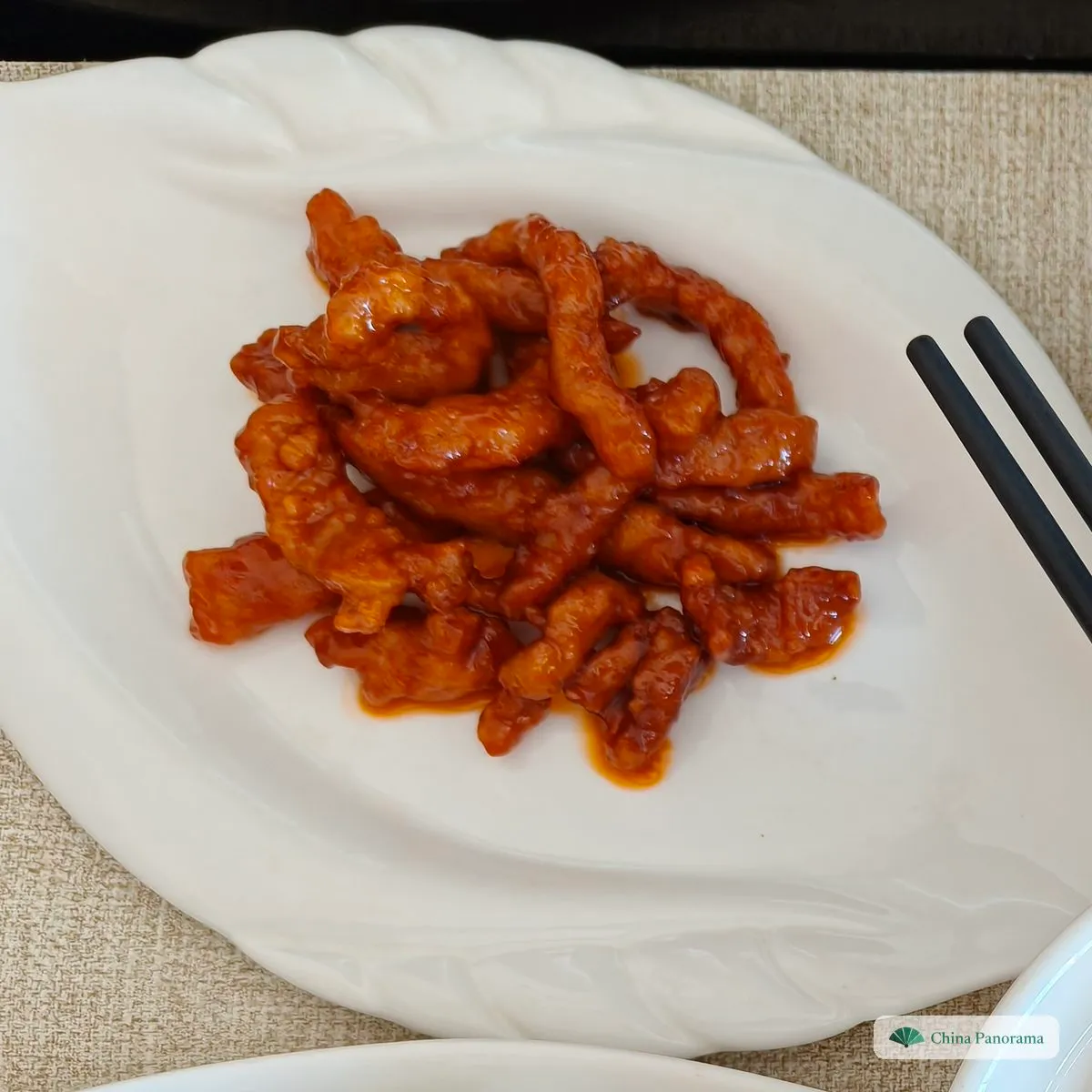 Thin strips of battered pork in a bright red-orange sauce on a leaf-shaped white plate, chopsticks on the right