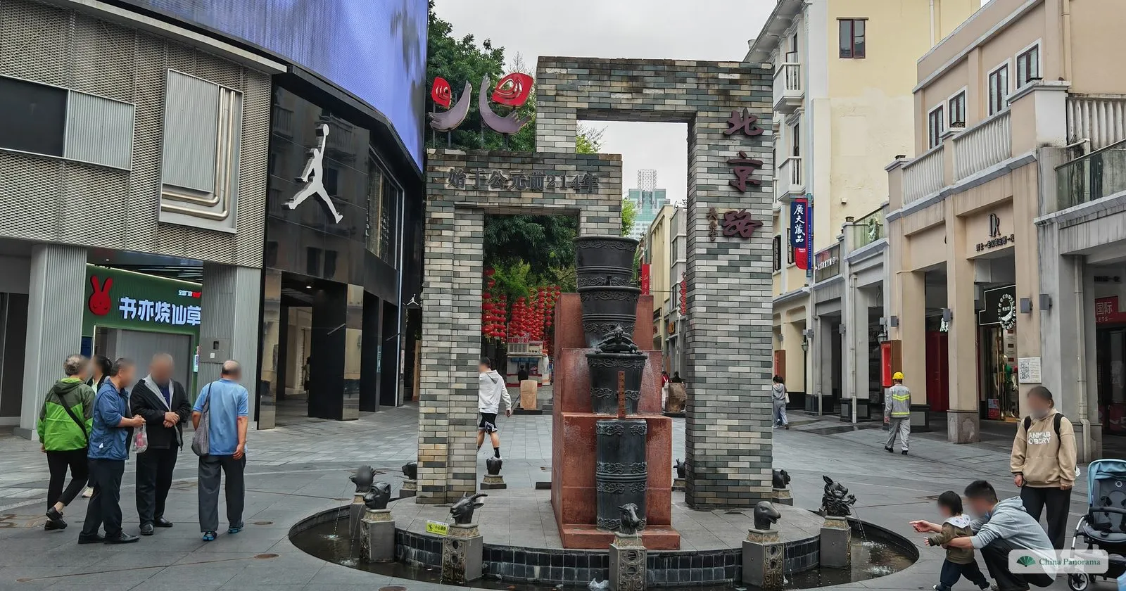 A grey-brick gateway sculpture with the characters 北京路 on a small fountain at Beijing Road, with bronze figures around the basin and passers-by