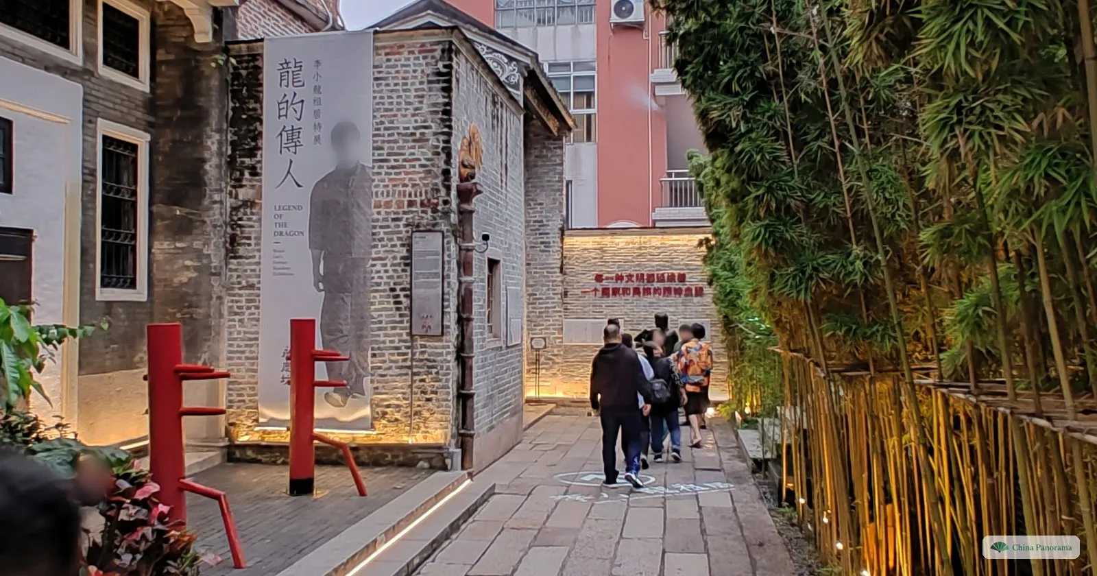 A narrow grey-brick lane lit from below, with red wooden wing chun training dummies, a tall banner showing Bruce Lee and visitors walking towards a lit wall