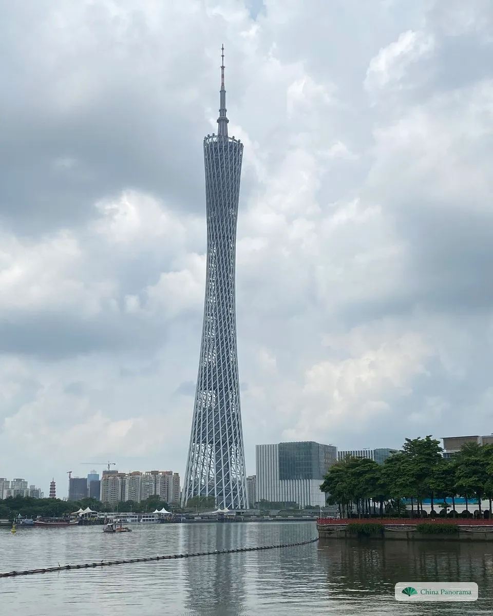 The Canton Tower by day across the Pearl River, its grey lattice body and mast rising above the riverbank under a cloudy sky