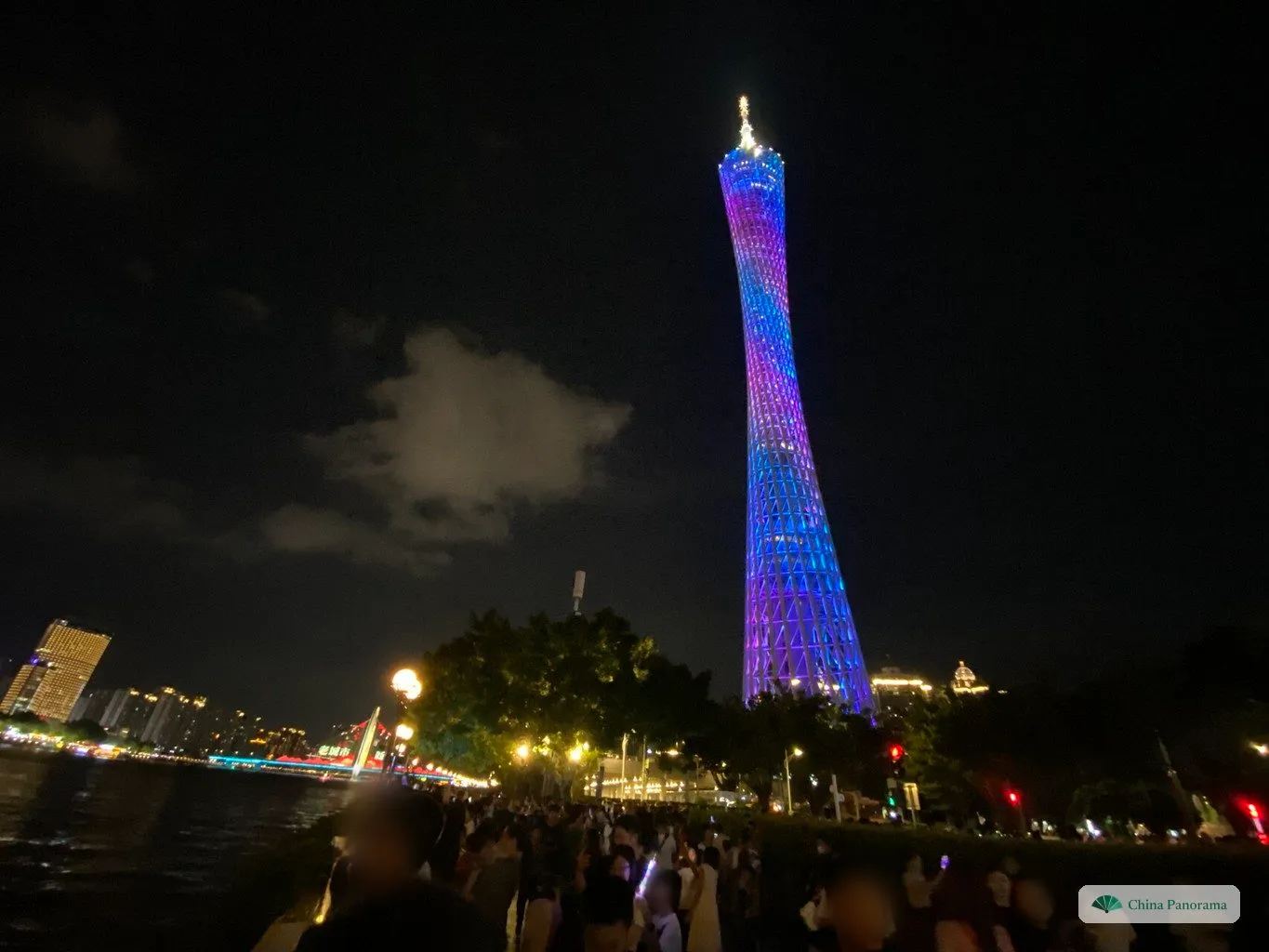 The Canton Tower lit in purple and blue at night, seen across the river from the north bank, with a crowd on the promenade in the foreground