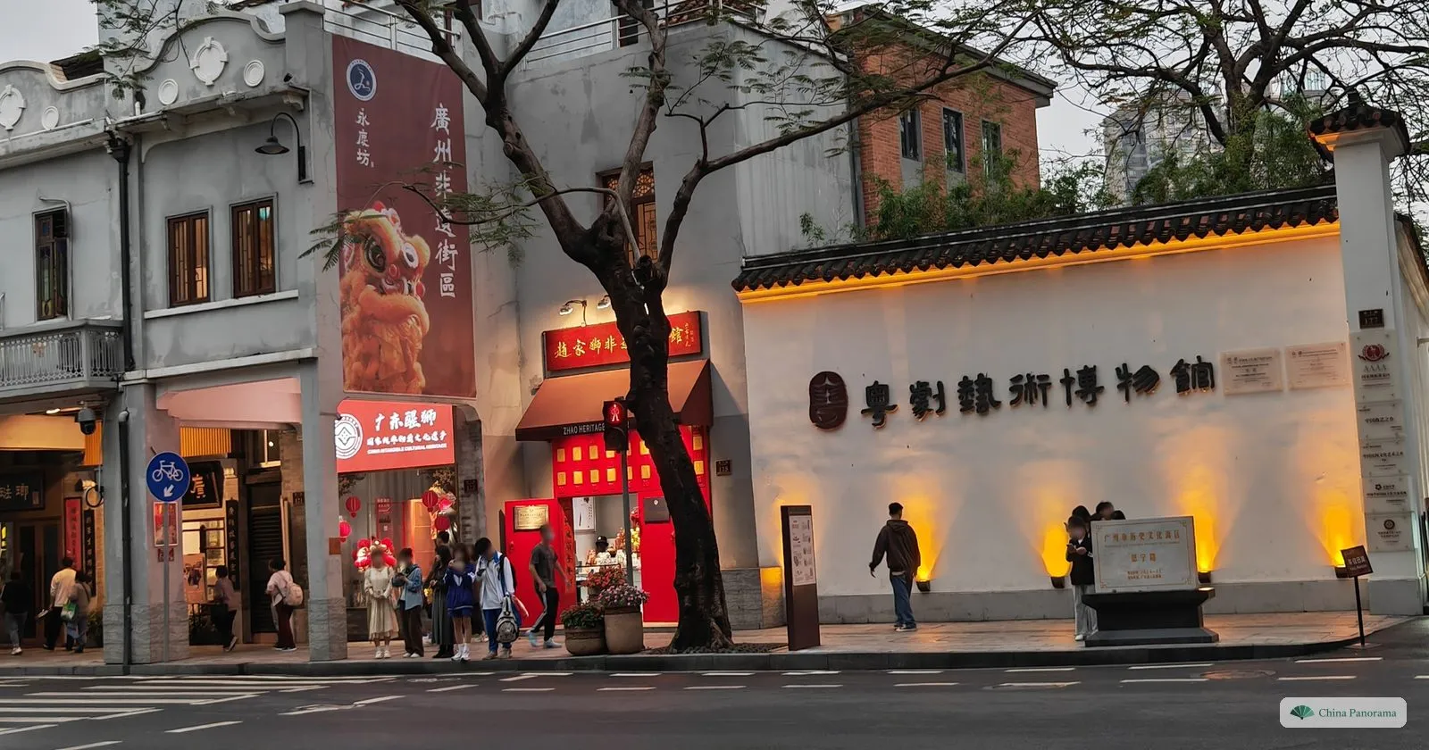 A white wall with the name of the Cantonese Opera Art Museum in dark characters, lit at dusk on Enning Road, next to arcaded shopfronts and a lion-dance banner