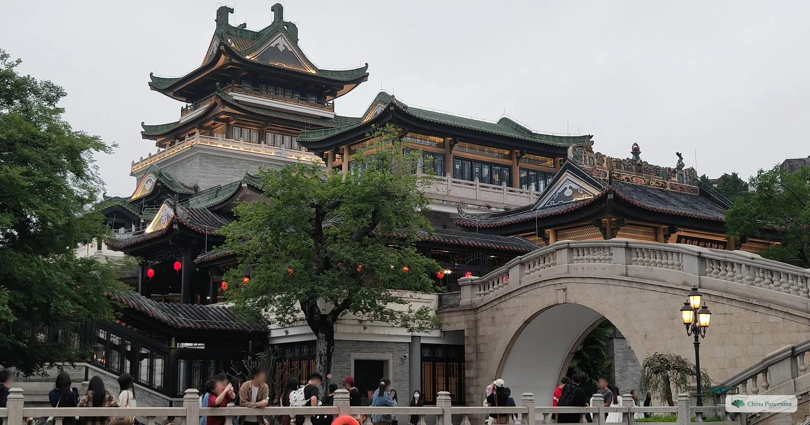 The Cantonese Opera Art Museum: grey-tiled halls and a tiered pavilion with upturned eaves behind a high white stone arched bridge, visitors at the railing in front