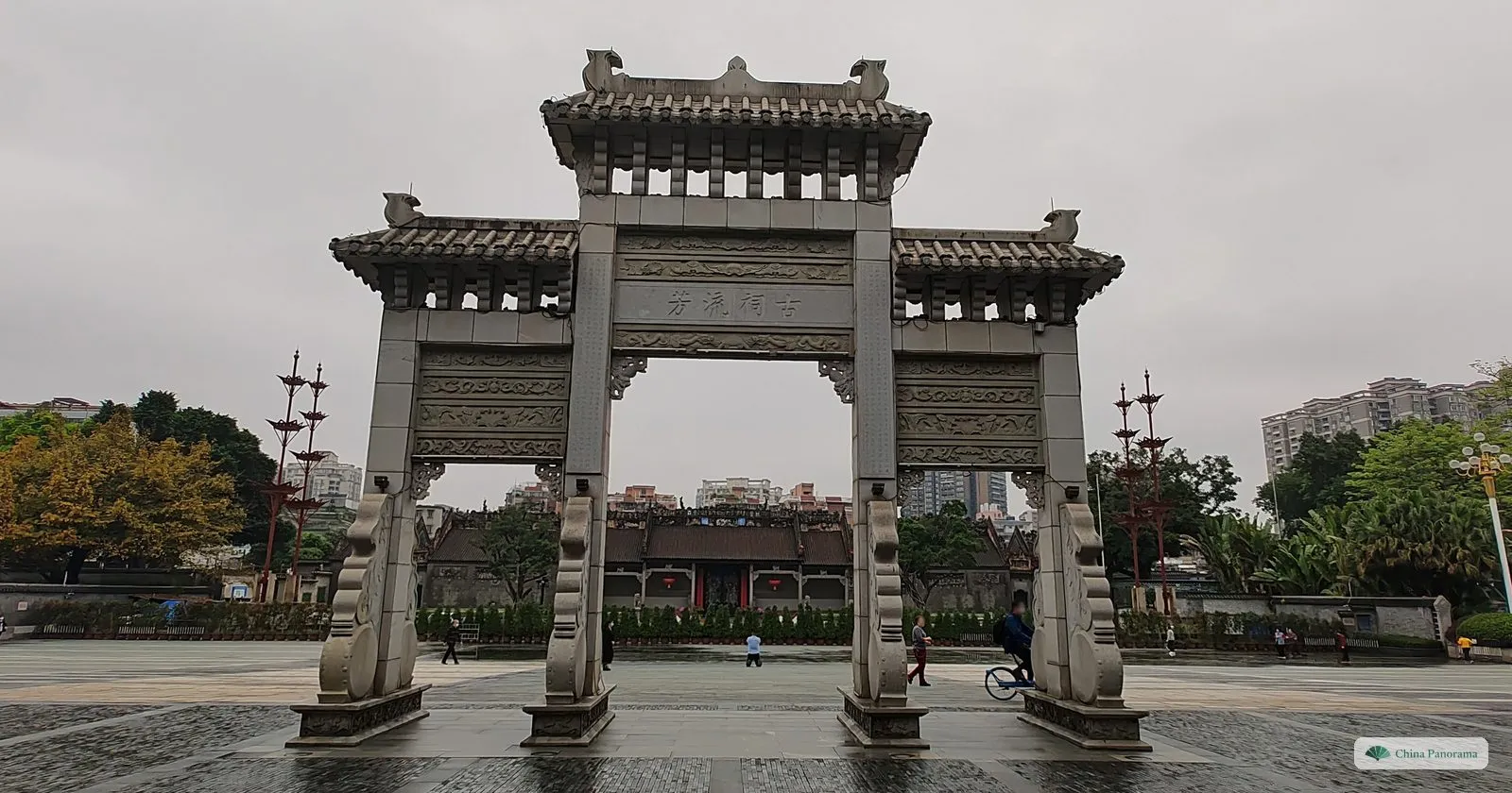 A granite memorial archway with three openings on the wet square in front of the Chen Clan Ancestral Hall, the low roofs of the hall behind it