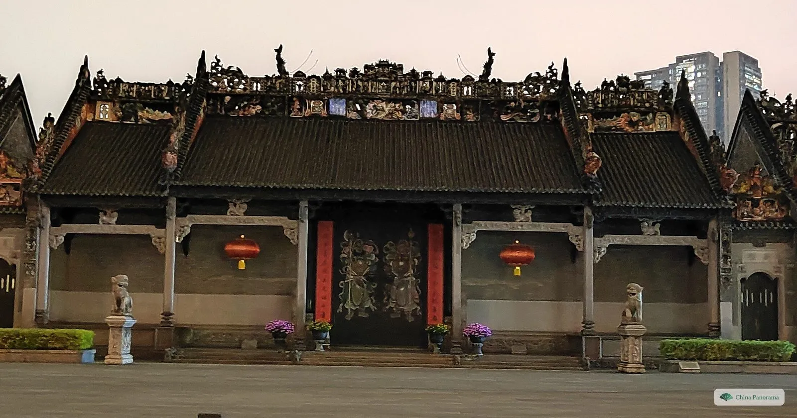 The front hall of the Chen Clan Ancestral Hall at dawn: a long grey-brick facade with granite columns, two red lanterns, painted door gods on the central doors, stone lions on pedestals, and a roof ridge crowded with ceramic figures against a pink sky