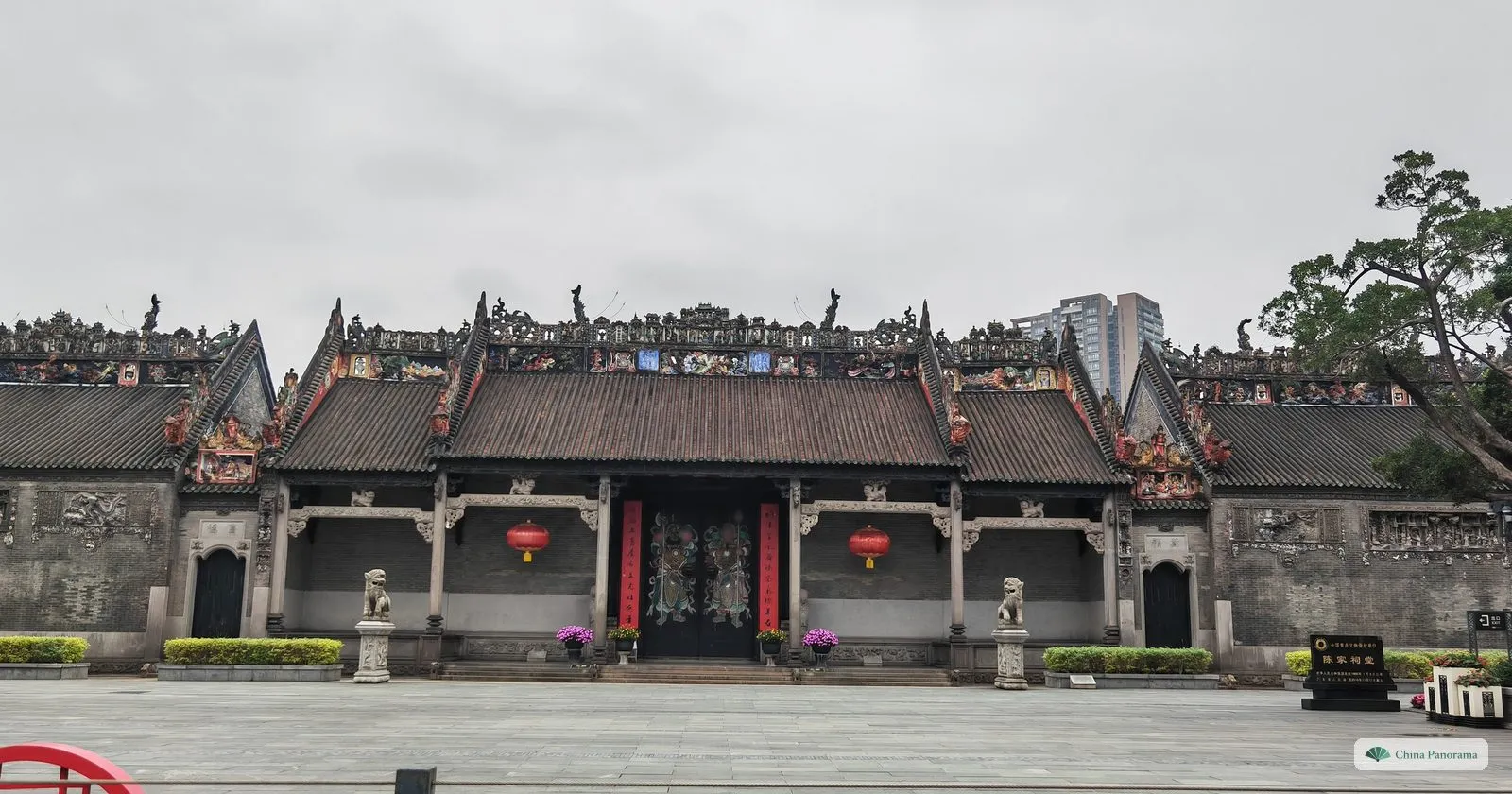 The whole front of the Chen Clan Ancestral Hall from the empty square on an overcast morning: three gabled roofs with ornate ridges, carved grey walls and a tree on the right