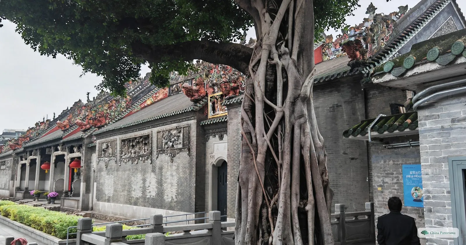 The side of the Chen Clan Ancestral Hall: grey brick walls with carved panels, roof ridges thick with colourful ceramic figures, and the hanging roots of a banyan tree in front