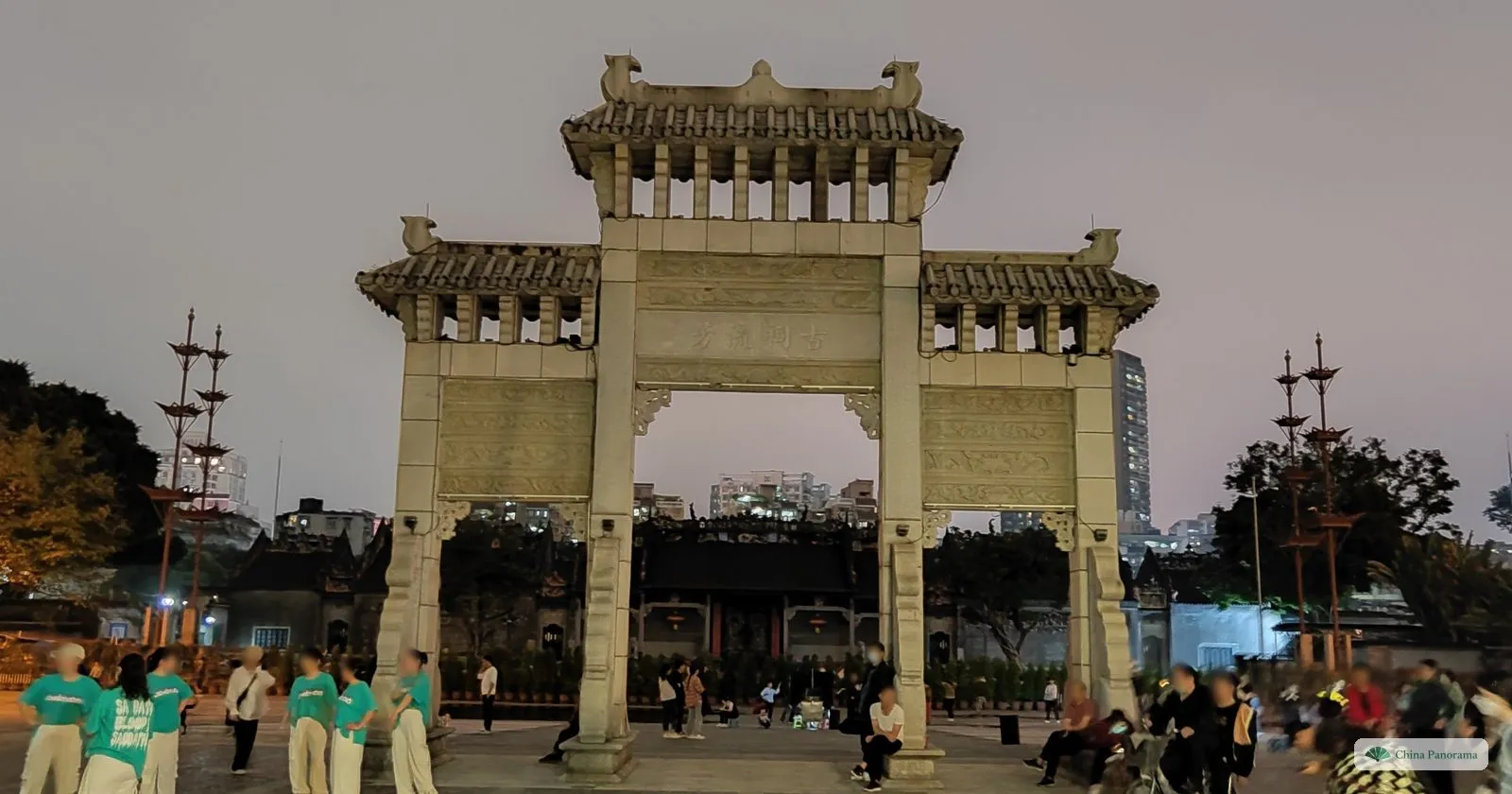 The granite archway on the Chen Clan square after dark, with a dance group in matching green T-shirts practising in front and people sitting on the steps