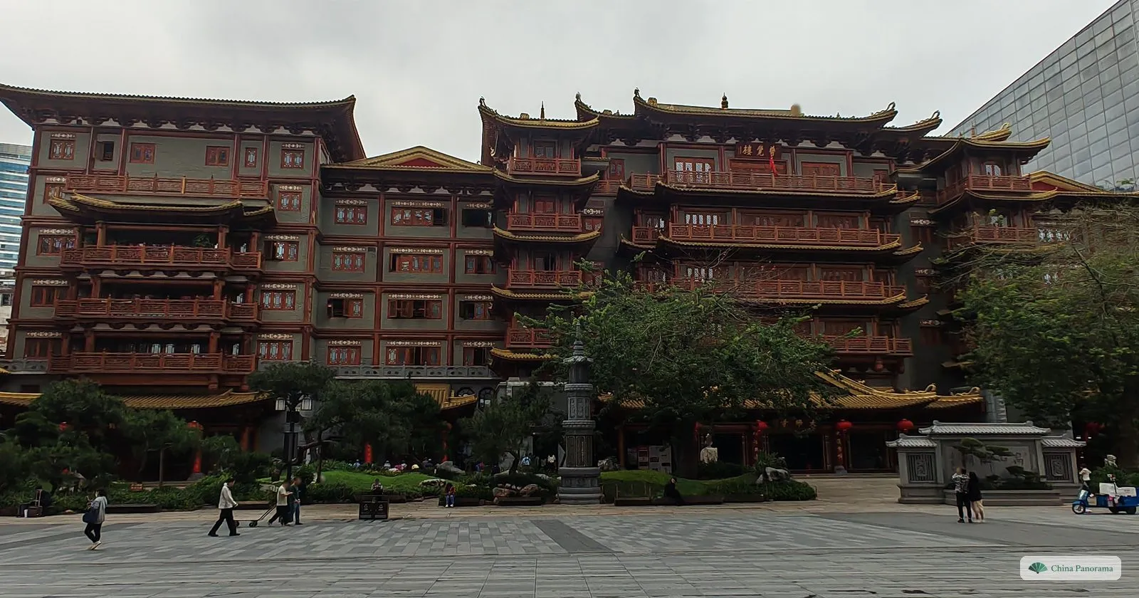 A huge new multi-storey temple building in red and gold with tiers of balconies, behind a paved square with a few visitors