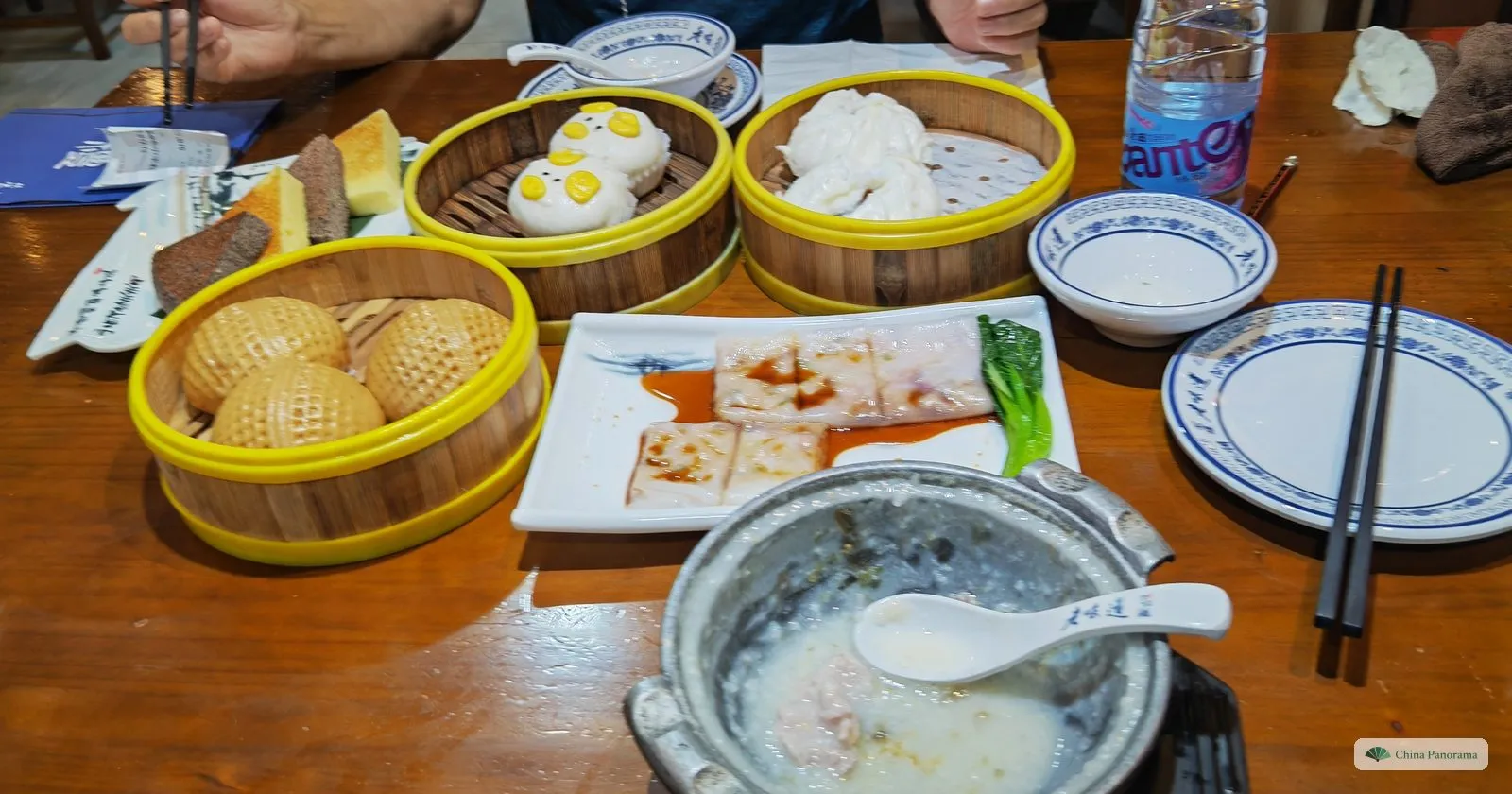 A Cantonese breakfast on a wooden table: bamboo steamers with buns, a plate of rice-noodle rolls with greens, congee in a clay pot and blue-and-white bowls