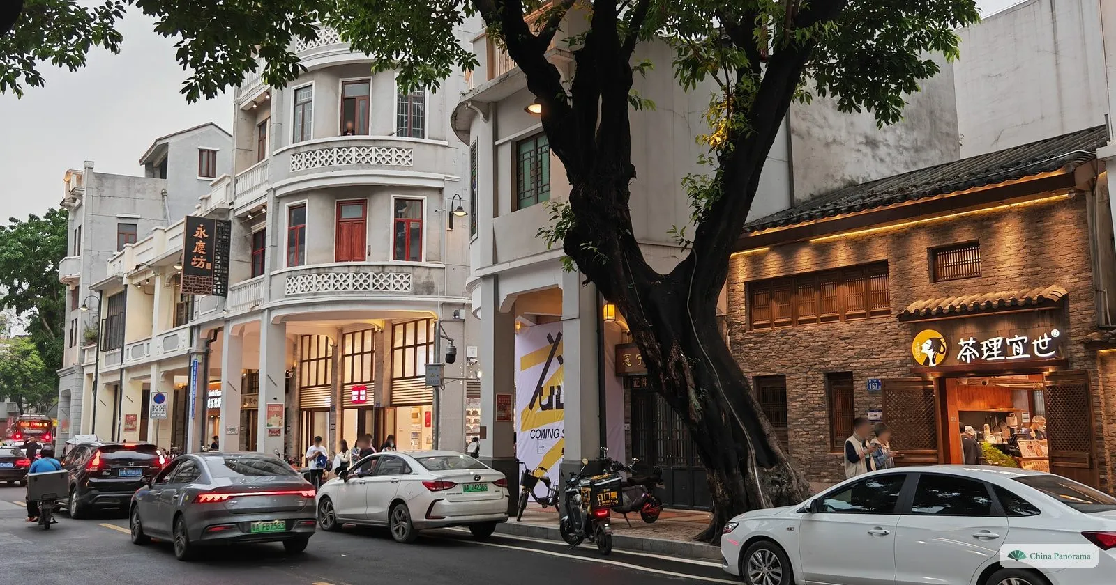 The pale arcaded shophouses of Enning Road at dusk, with a curved corner building, shops lit on the ground floor and cars parked along the street
