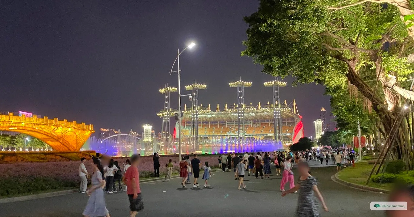 Haixinsha at night: a lit grandstand behind fountains, a golden arch structure on the left and families walking along a path lined with flower beds