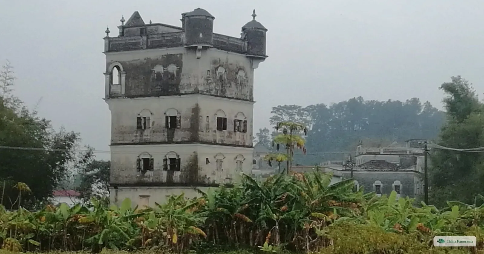 A weathered concrete diaolou tower with corner turrets and small shuttered windows, rising above banana plants in drizzle, with another tower behind