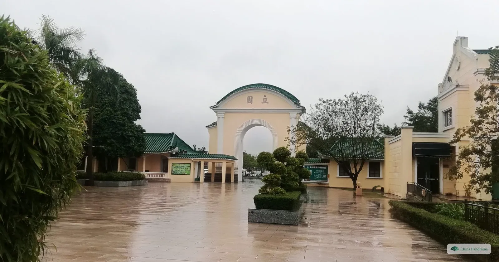 A cream-coloured triumphal arch with the name 立園 in a wet garden courtyard with clipped shrubs and green-tiled pavilions
